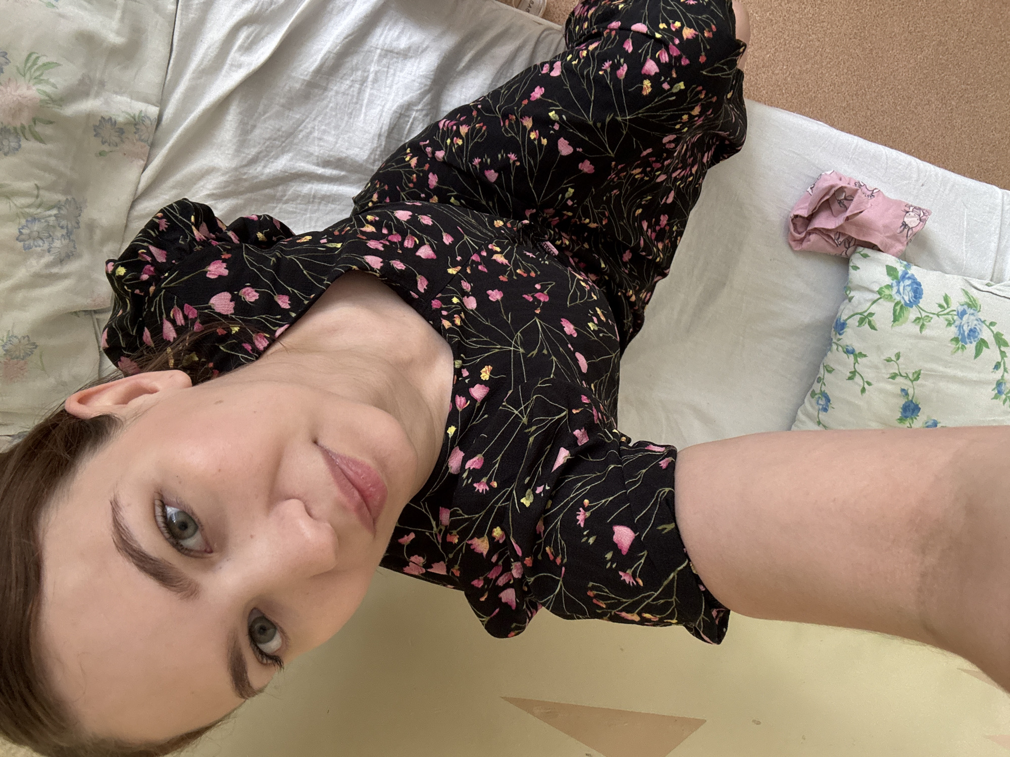 A young woman with light skin and brown hair is lying on a bed. She is wearing a black dress with a floral pattern of pink and yellow flowers. The bed has white sheets and a pillow with a blue floral pattern. There is also a pink scarf on the bed. The room appears to be a bedroom, with a yellow wall in the background. The image was taken indoors. The specific location in Chaikovsky, Russia is not discernible from the image.