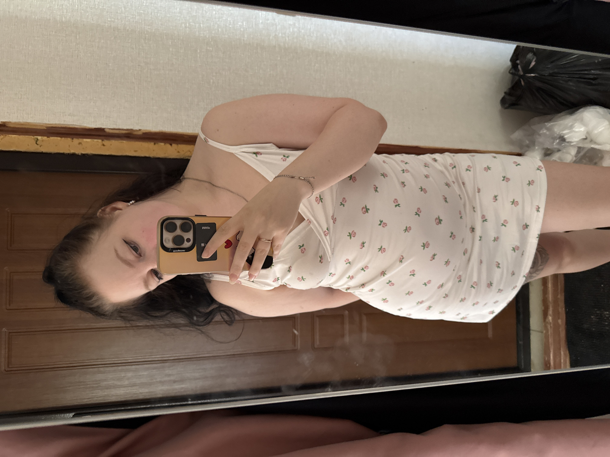 A young woman with dark hair pulled into a ponytail is taking a selfie in a mirror. She is wearing a white slip dress with a floral print. She is holding a smartphone in her right hand, and a ring is visible on her right ring finger. A silver bracelet is on her left wrist. The background appears to be an interior setting with a wooden door and a textured wall. A large mirror is positioned to her left, reflecting her. To her right, there are two trash bags. The image appears to have been taken in the city of Vylgort, Russia.