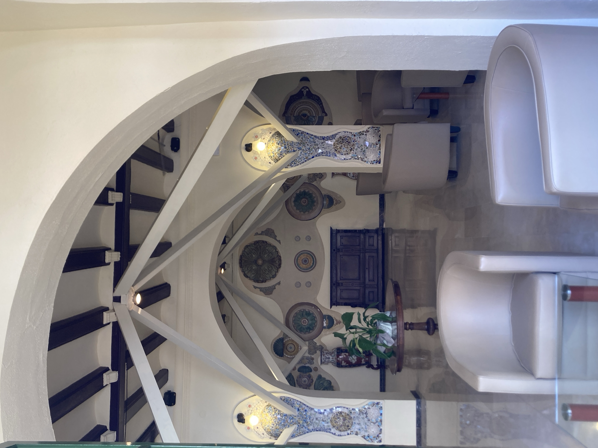 This image captures the interior of a lobby, likely in Lloret De Mar, Spain, with a focus on its architectural and decorative elements. The scene is dominated by a series of curved arches and angular beams that create a dynamic ceiling structure, reminiscent of a vaulted or tent-like design. Dark wooden beams are visible within the white ceiling, adding contrast.
The walls are adorned with colorful mosaic panels shaped like elongated urns or stylized figures, flanking ornate circular wall decorations that resemble medallions or rosettes in shades of green, brown, and blue. In the center of the background, a dark wooden cabinet with intricate detailing serves as a focal point. A round wooden table with a potted plant and a vase sits in front of the cabinet, reflecting on the polished marble floor.
In the foreground and mid-ground, several light-colored, upholstered armchairs are arranged, suggesting a waiting or seating area. One armchair is prominently featured in the lower right of the frame, while others are visible further back. The lighting appears to be a mix of ambient overhead lights and decorative lamps integrated into the mosaic columns. The overall impression is one of a stylish and possibly vintage or uniquely designed interior space. There are no people visible in the image.