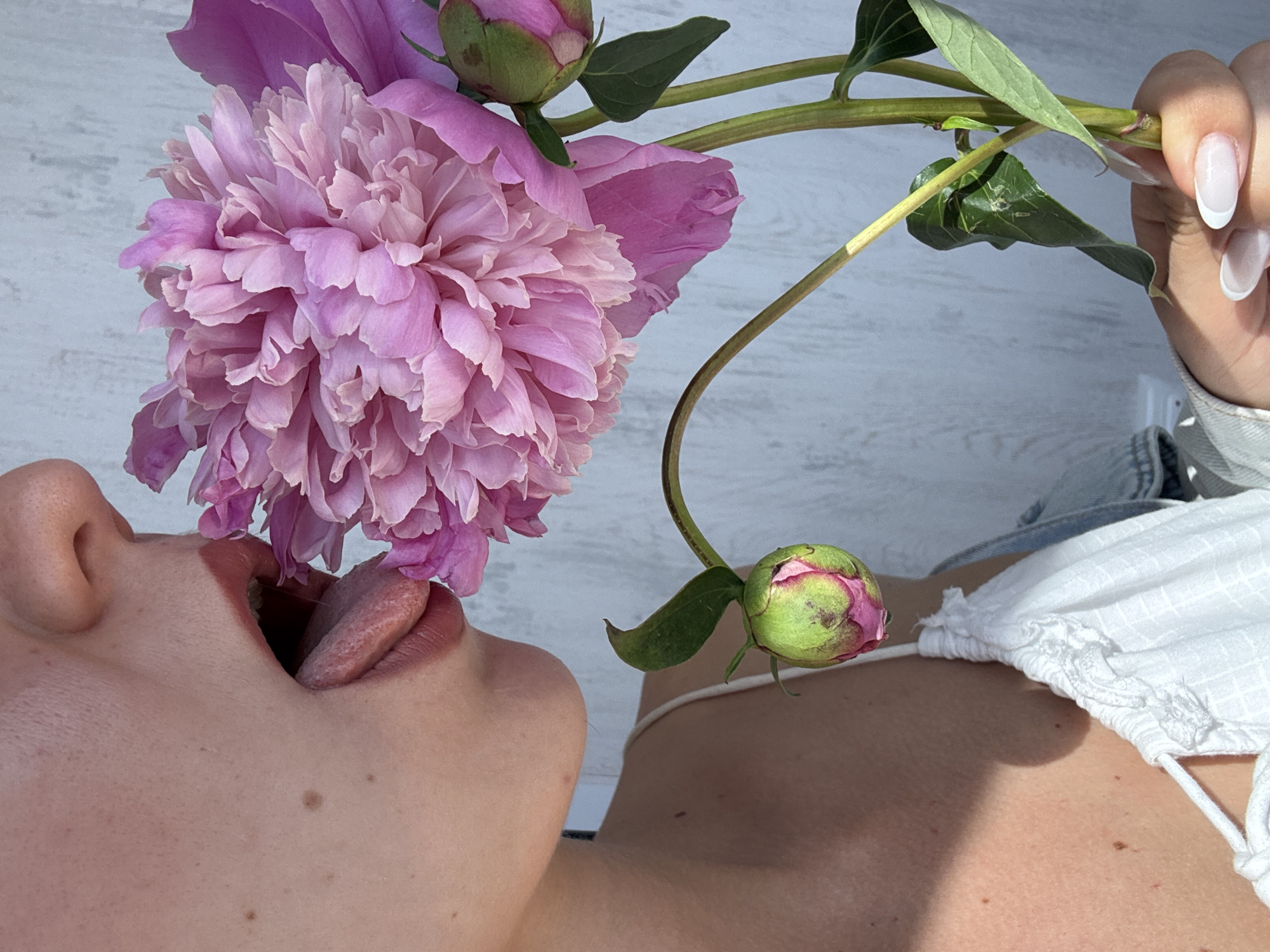 A close-up, eye-level shot shows a person's mouth with their tongue extended to touch a large pink peony flower. The flower is in focus, with detailed petals and two buds. The person's face is partially visible, showing skin with freckles and lips with lipstick. They are wearing a white camisole. The background is a light-colored, textured wall. The lighting suggests an outdoor setting, possibly during the day. The person's hand, with painted fingernails, is holding the stem of the flower.
