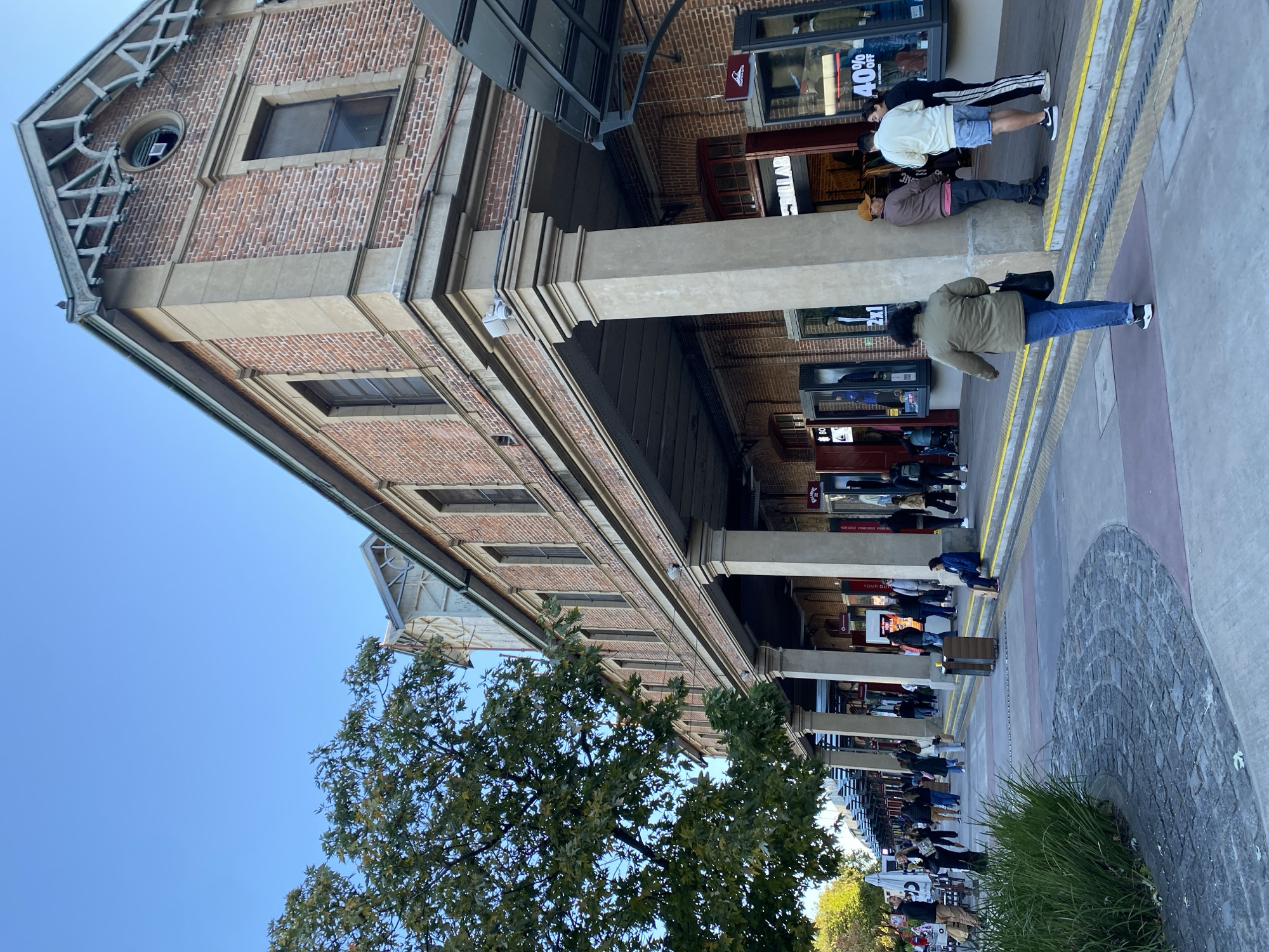 The image depicts a partially shaded street scene in Buenos Aires, Argentina, with a long brick building dominating the left side of the frame. The building features rows of windows and a distinct architectural design at the top. Large, rectangular concrete pillars support an overhang that shelters the sidewalk in front of the building. People are walking along the sidewalk, some appearing to be shopping, with storefronts visible behind them. A tree is visible in the foreground on the left, partially obscuring the building. The sky is clear and blue. The setting appears to be a commercial area with pedestrian traffic. A low stone wall or landscaping feature is visible on the right side of the image.