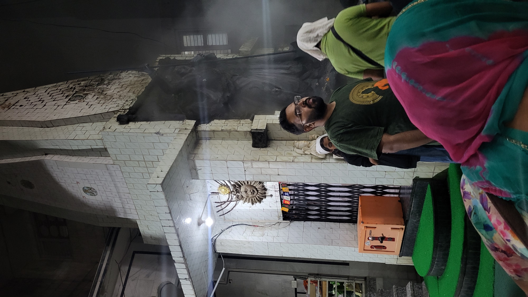 The image depicts an indoor scene, likely within a spiritual or religious establishment in Kasol, India. The space is characterized by white tiled walls, some of which show signs of discoloration or wear.
Several people are visible, gathered in what appears to be a queue or a viewing area. In the mid-ground, a man with a beard and glasses, wearing a dark green t-shirt with a golden circular design, is looking upwards and to his left. Behind him and to his right, another individual wearing a light green shirt and a white head covering is seen from the back. On the far right, partially cut off by the frame, is a person wearing a vibrant pink, blue, and white patterned garment, seen from behind. Another person in a white head covering is partially visible behind the man in glasses.
A prominent decorative feature on a wall comprises a sun-like face with radiating elements, adorned with a small golden disc. A bright light bulb hangs near this feature. Below it, a black metal shutter or gate is partially open. To the right of the shutter, an orange metal box or safe with a handle and lock is placed on green carpeted steps that ascend towards the right side of the image. Faint text, possibly "TUL" or "JUL" and "LOCK," can be seen on the orange box.
The upper background is filled with a significant amount of smoke or mist, which obscures large, dark-colored stone or concrete structures and a barred window. This haze suggests an ongoing ritual involving incense or fire. The dim lighting implies it is either evening or night, with artificial lights providing illumination to the scene. The overall ambiance is one of observation and reverence, typical of a place of worship or spiritual significance.