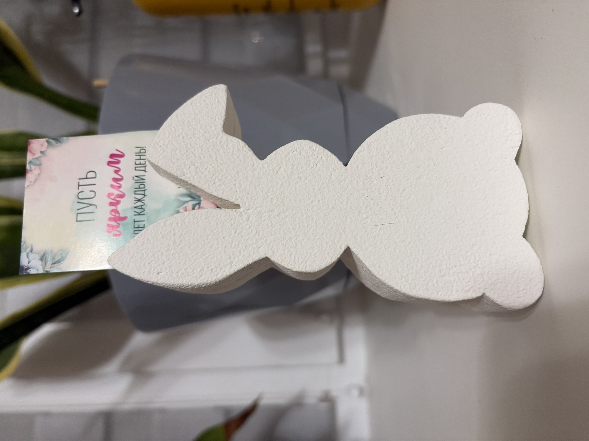 A close-up shot shows a white, decorative bunny figurine with a textured surface, lying on its side. A card with Russian text ("Пусть Чудим Ждет каждый день!") is partially visible behind the bunny. The objects are positioned on a light-colored surface. A grey container and green plant leaves are visible in the background.