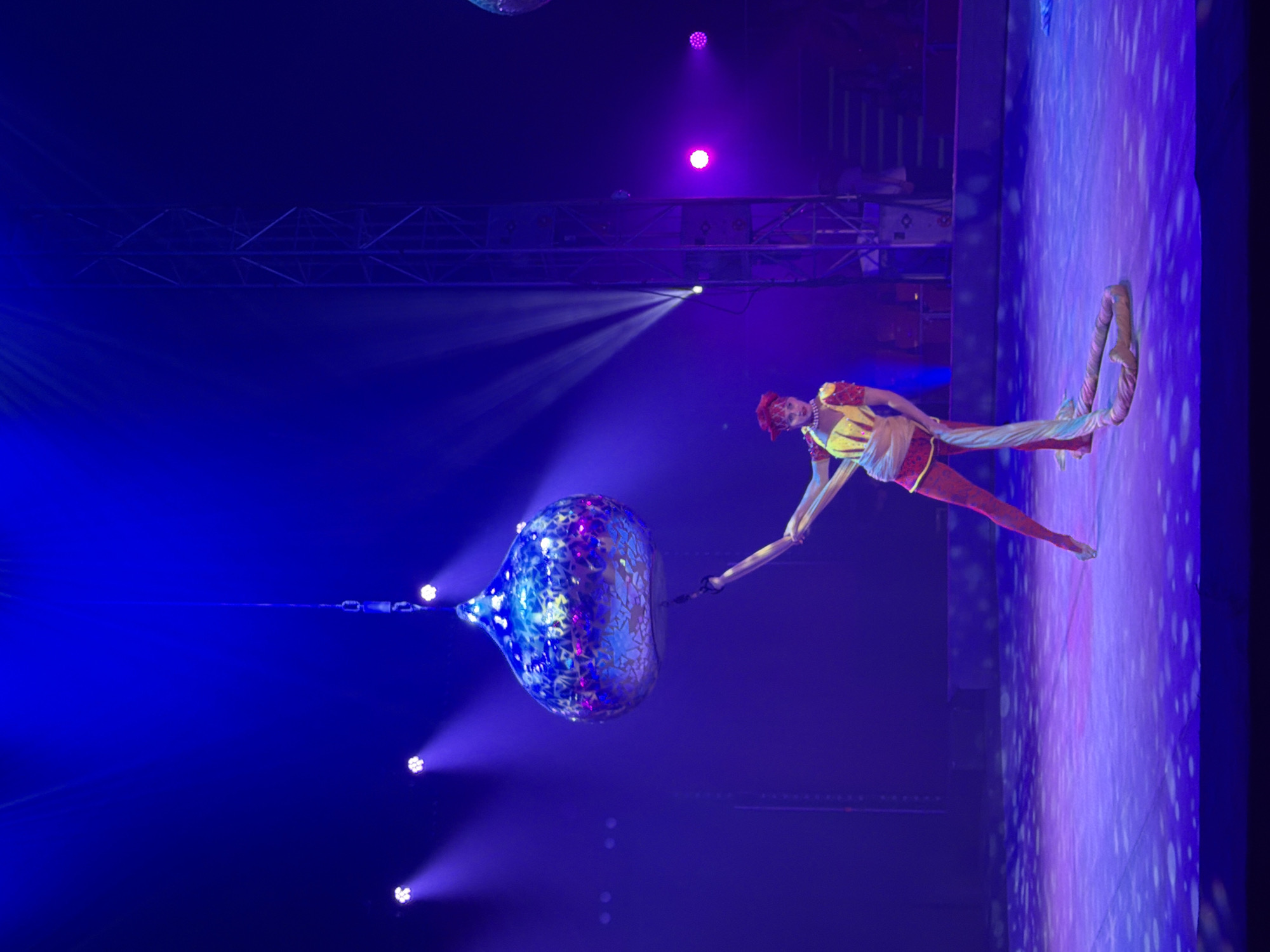 This media file shows an aerial performer in a circus or stage setting. The performer, dressed in a vibrant yellow and red costume with a red headpiece, is suspended in mid-air. They are elegantly positioned, with one leg extended and their arm reaching towards a large, spherical, iridescent object that hangs from above. The object is adorned with a reflective, patterned surface that catches the light, displaying shades of blue, purple, and pink.
The background is dark and atmospheric, illuminated by dramatic stage lighting. Several bright spotlights cast beams of light from above and the side, creating a theatrical effect. There are also purple lights emanating from higher up, adding to the visual spectacle. The floor or stage area is covered in a projected pattern of light, creating a dynamic and colorful surface that reflects the lighting from above. The overall scene suggests a performance taking place at night, likely indoors in a venue designed for such displays, such as a theater or circus tent. The performer's posture and the grand scale of the props indicate a moment of artistic expression and skill, possibly a dance or a balancing act. There is no visible text in the media file.