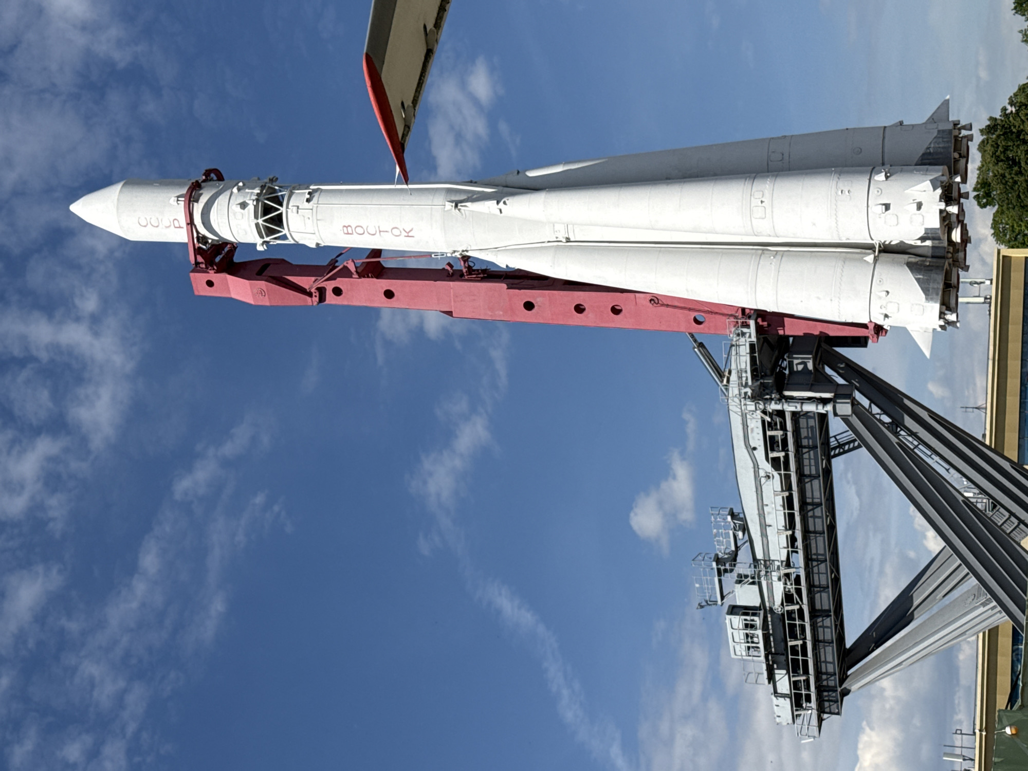 A large, white Vostok rocket is prominently displayed horizontally against a clear, bright blue sky with scattered white clouds. The rocket is held aloft by a sturdy red truss support structure or cradle. On its conical nose fairing, the Cyrillic letters 'CCCP' are visible in red, signifying its Soviet heritage. Below this, along the central section of the rocket's body, the name 'BOCTOK' (Vostok) is also clearly printed in red Cyrillic.
The rocket exhibits a multi-stage configuration, characteristic of the R-7 family. Four distinct, cylindrical booster stages flank a central core, each terminating in a cluster of engine bells at the rear. The rocket's primary color is white, creating a stark contrast with the red support structure and the red text.
To the far right of the frame, a large, dark grey metallic gantry or industrial structure is partially visible, suggesting a permanent outdoor exhibit or a museum setting. Lush green tree foliage also peeks into the frame from the upper right, adding a natural element. The scene is illuminated by daylight, under what appears to be pleasant weather. Given the rocket type and the location hint of Khimki, Russia, this is likely a monument or exhibit celebrating the historic Soviet Vostok space program.