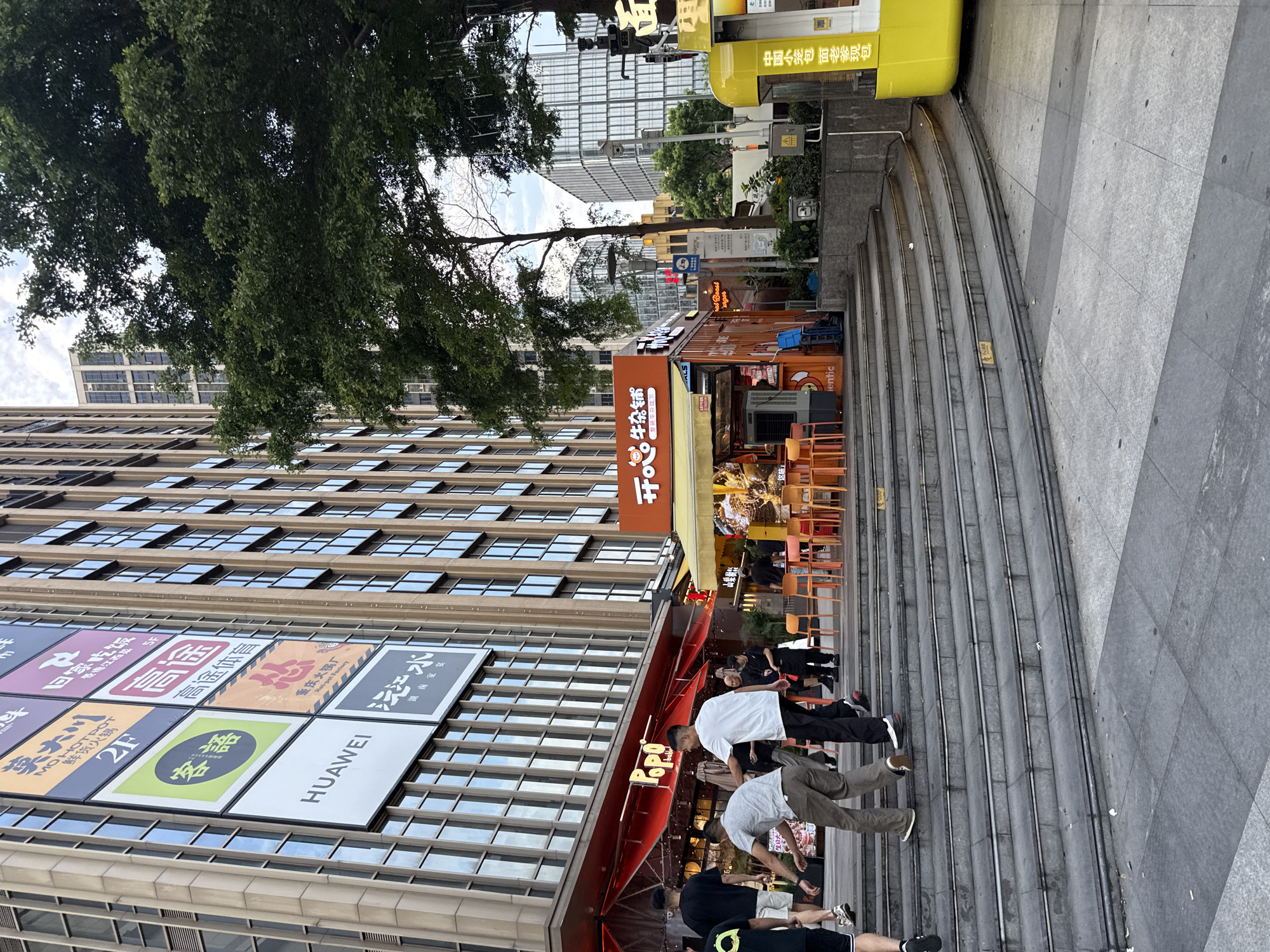 In Haizhu District, China, a wide set of concrete stairs leads up to a bustling street scene. On the left, a large building displays numerous colorful advertisements, including one for HUAWEI. To the right of the stairs, an orange food kiosk with outdoor seating is visible, with patrons and staff around. Further to the right, a bright yellow kiosk stands, likely a vendor. Several individuals are captured in motion on the stairs and in the vicinity of the kiosks, suggesting a lively urban environment. The background features more contemporary buildings and lush green trees, indicating a blend of modern architecture and natural elements. The lighting suggests it is daytime.