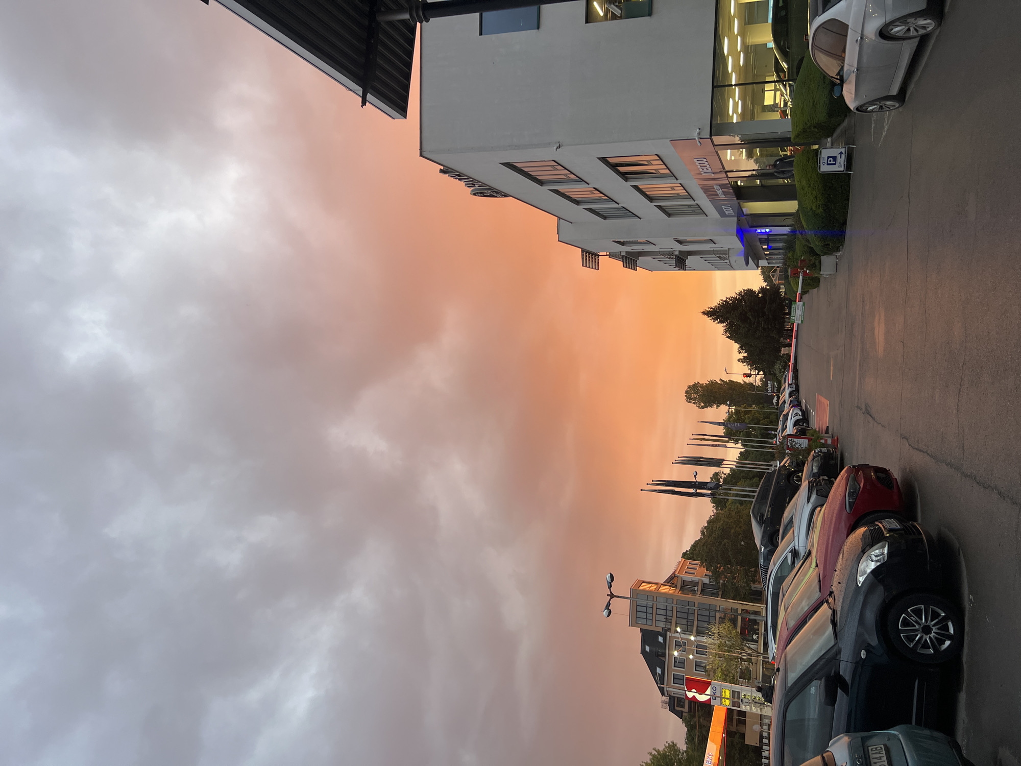 Captured from ground level in the city of Sofia, Bulgaria, the image displays a sky transitioning from grey, overcast clouds to a vibrant orange hue, indicative of either sunrise or sunset. The foreground features a row of parked cars along a paved road or parking lot, with a multistory building to the right. Street lights are visible among the buildings, and a few trees are also present. A commercial or office building, partially visible, is situated on the left, with signage displayed on its facade. The lighting suggests it may be dusk. No people are discernible in the image.