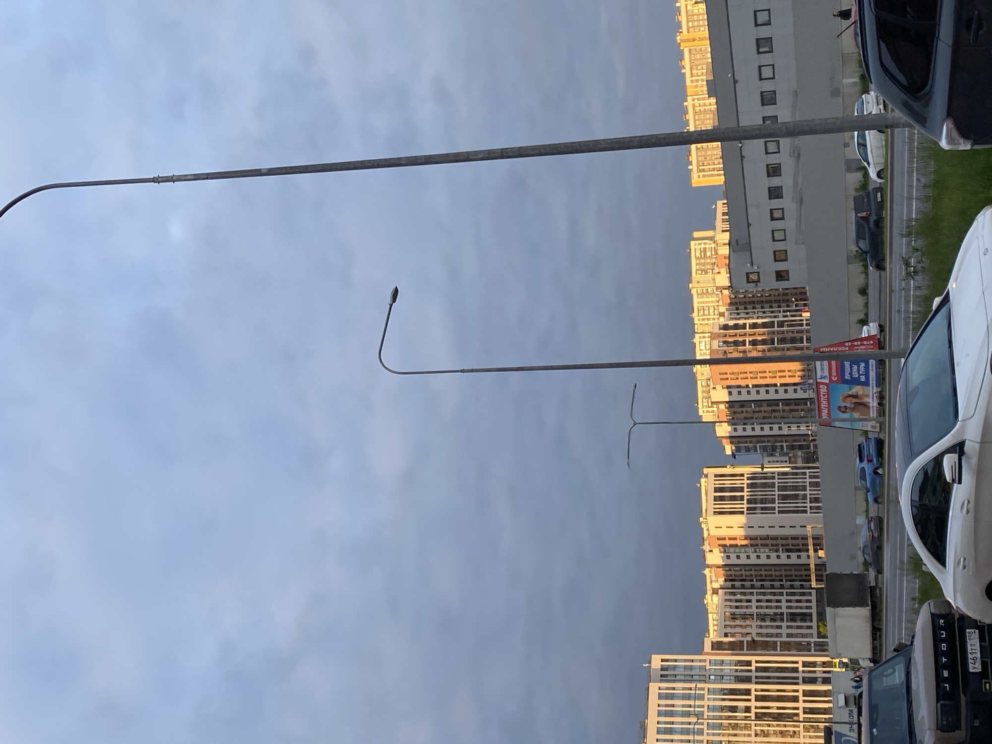 This image captures a modern urban landscape in Saint Petersburg, Russia, under an overcast sky, likely during the late afternoon or early evening as suggested by the sunlight catching the upper floors of buildings. The scene is dominated by several multi-story residential buildings, mostly on the right side, featuring contemporary architecture with numerous windows.
In the foreground and middle ground, several streetlights are prominent. One long, straight lamp post extends horizontally across the upper part of the frame, while another features a distinct curved arm with a light fixture. A third T-shaped streetlight is also visible. The sky is predominantly light grey and cloudy, hinting at overcast weather.
Parallel to the buildings runs a road or street, bordered by some green space. Various vehicles are present, including a prominent white van or SUV in the immediate right foreground, and a dark grey or black SUV with "JETOUR" visible on its rear, which also displays a Russian license plate "y461TT 198" (with 198 being a Saint Petersburg region code). Other cars, both white and dark-colored, are visible parked along the road.
A billboard or advertisement stand is positioned near the buildings, displaying Cyrillic text. Visible words include "РЕКЛАМА" (Advertisement), "ЛУЧШИЕ ЦЕНЫ НА ТУРЫ" (Best prices on tours), and "ПРАВИТЕЛЬСТВО" (Government). The overall impression is a typical city street scene, with no specific activity or people clearly discernible.