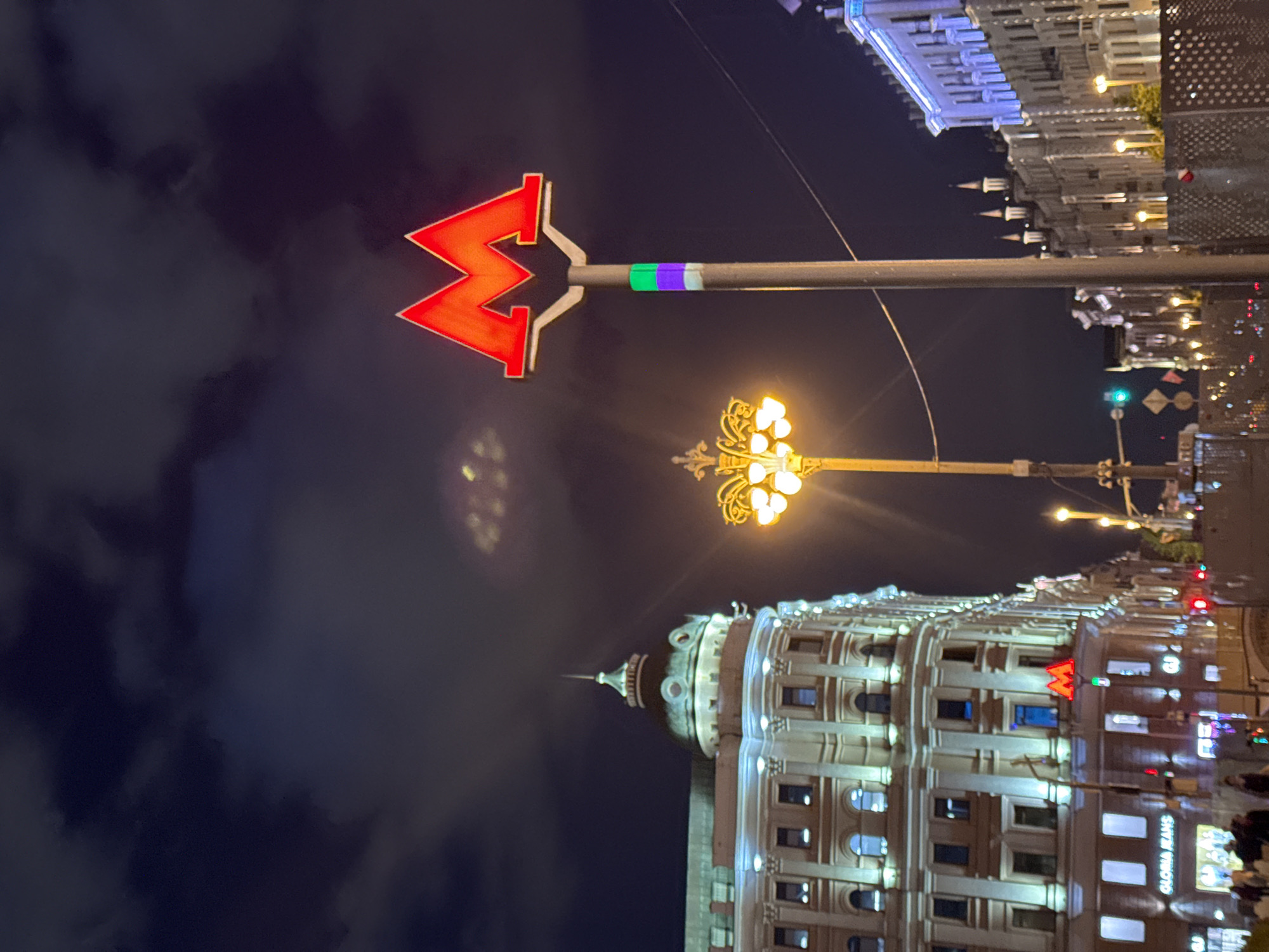 This media file captures an illuminated urban street scene at night in Moscow, Russia. A prominent, bright red "M" logo, symbolizing a Moscow Metro entrance, is affixed to a tall pole in the foreground. The pole beneath the "M" features distinct green and purple bands.
To the right of the metro sign, a classical streetlamp with multiple ornate, glowing yellow bulbs casts light on the surroundings. In the background, several grand, multi-story buildings with intricate architectural details line the street. Their facades are brightly lit, highlighting numerous windows, some of which glow from within. One building on the right side features a distinctive dome and spire.
The dark night sky is visible above, with some faint cloud formations that reflect the city's ambient light. Overhead wires are also discernible, crisscrossing the upper portion of the frame. Blurry figures near the bottom right suggest the presence of pedestrians, and distant lights hint at ongoing traffic or other urban activity. Visible text on one of the buildings reads "GLORIA JEANS." The overall atmosphere is that of a bustling and majestic city during the evening hours.