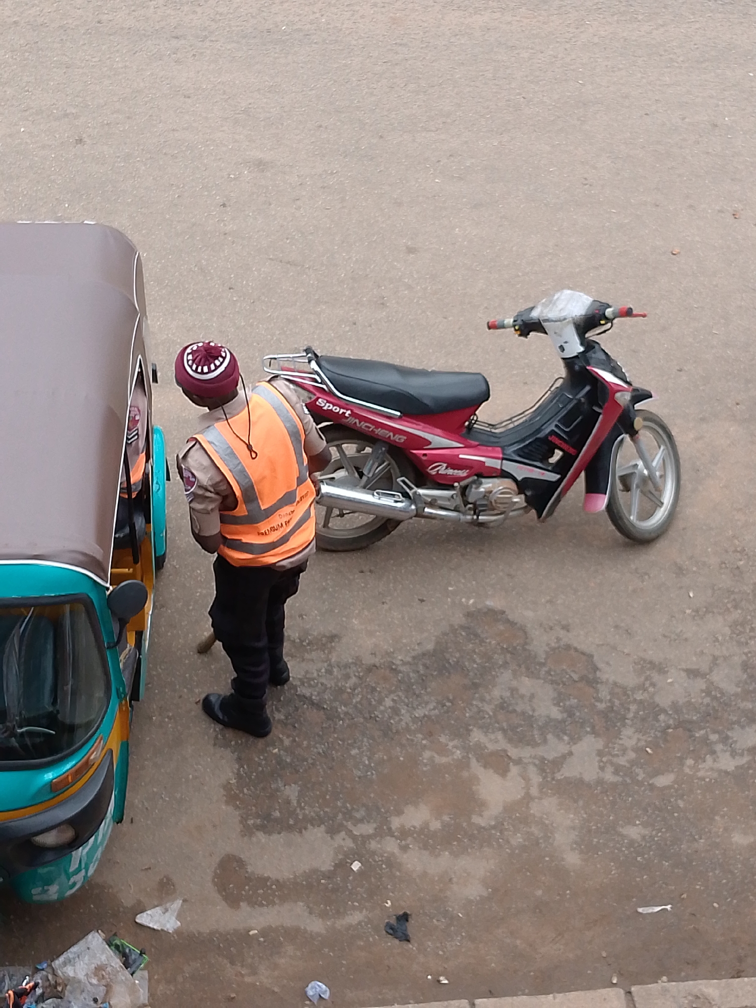 In Bukuru, Nigeria, a traffic warden wearing a maroon and white patterned cap, a tan uniform, and an orange high-visibility vest stands next to a green and brown auto-rickshaw. The warden is looking down at something on the ground, holding a broom. To the right of the warden and auto-rickshaw is a red and black motorcycle. The ground is paved with asphalt and appears damp in places. There is some debris scattered on the ground near the auto-rickshaw, including plastic bags and what appears to be trash.