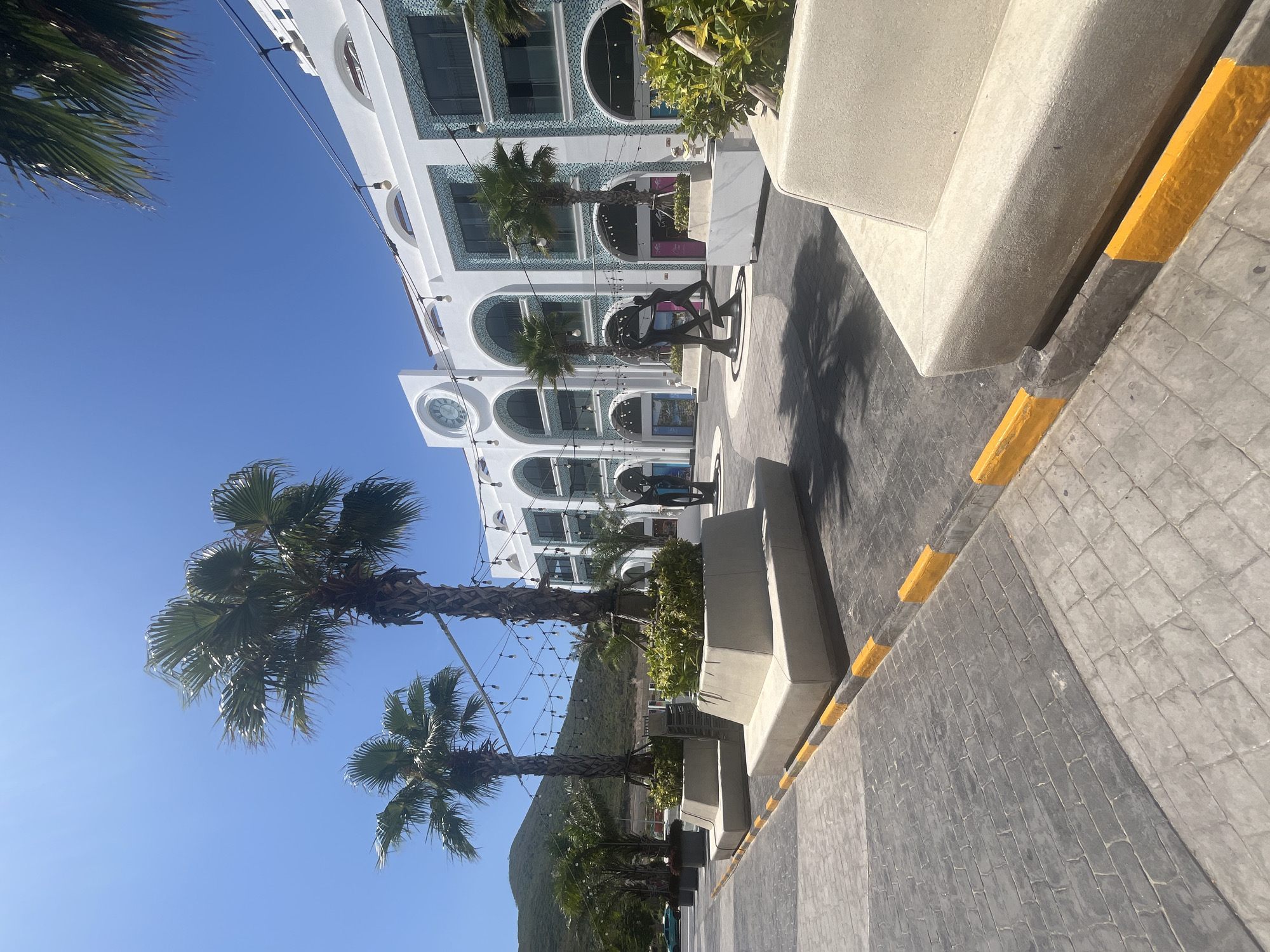 The image shows an outdoor urban scene, likely a pedestrian area or walkway in the city of Unknown, Unknown. The ground is paved with grey bricks and a curb that is yellow. On the side of the walkway are planters. In the background is a white building. Palm trees are visible along the walkway and in front of the building. In the background is a mountain covered in trees.