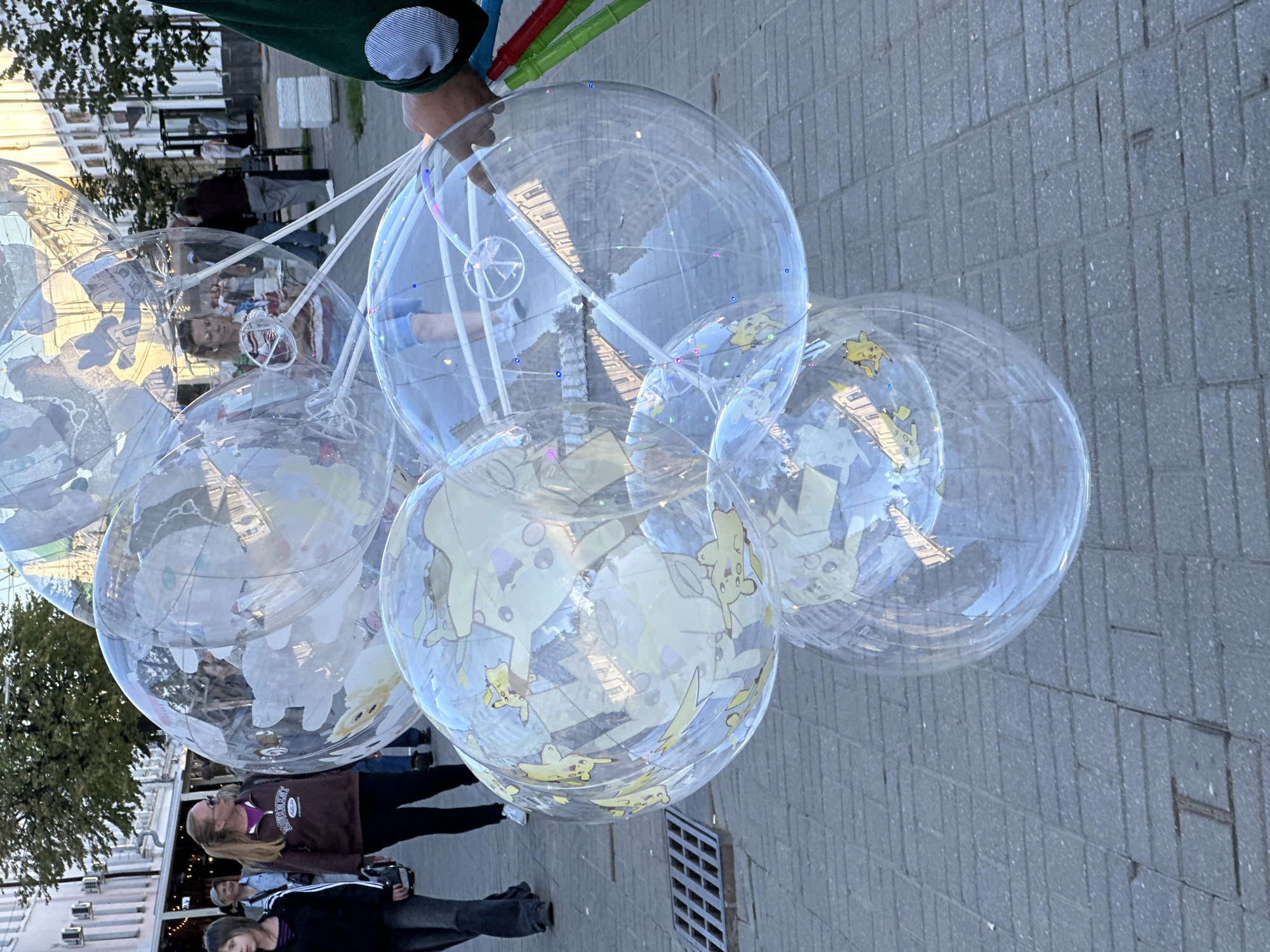 The image depicts a cluster of large, transparent inflatable globes, some adorned with images of Pikachu. They are held aloft by unseen strings attached to a set of wands, one of which is partially visible in the lower right corner being held by a person. In the background, several individuals are walking on a paved walkway. The setting appears to be an outdoor urban environment, possibly a street or plaza, with buildings and trees visible in the distance. The ground is paved with interlocking grey bricks. This scene is located in the city of Feklino, Russia.