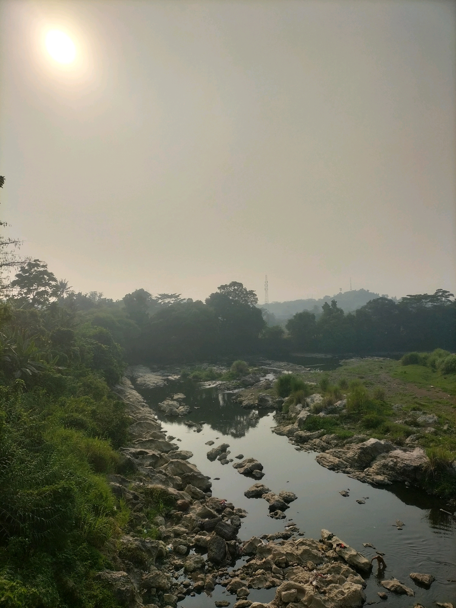 The image depicts a natural scene with a river flowing through a rocky, vegetated area under a hazy sky with a bright sun. Lush green vegetation lines the left bank of the river, which is characterized by numerous large, light-colored rocks scattered along its edges and within its shallow waters. The river itself appears dark and somewhat murky. In the background, a dense line of trees forms a natural barrier, with hills and some man-made structures, possibly communication towers, visible in the distance through the atmospheric haze. The overall impression is one of a somewhat wild and undeveloped landscape, possibly on the outskirts of a city given the distant structures.