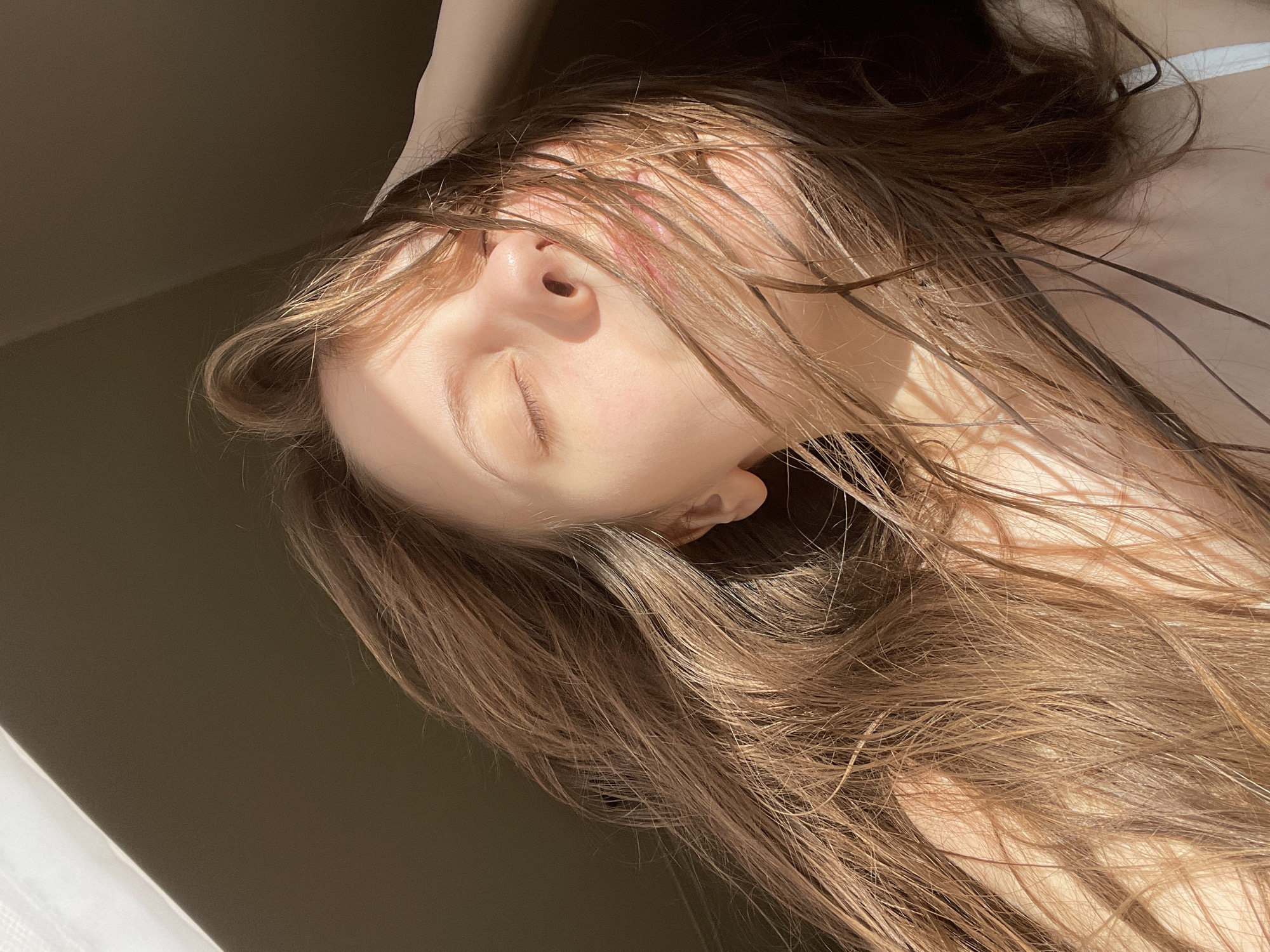 The image shows a close-up, angled shot of a person's face and long, brown hair, illuminated by a strong, warm light that casts shadows. The person is lying down, with their eyes closed, and their head is tilted back, with one hand resting on their head. The light creates a dappled effect on their skin and hair, highlighting strands and creating a soft glow. The background is mostly dark, with a diagonal line of lighter color suggesting a wall or a surface. The overall mood is serene and peaceful, with a sense of natural light and quiet contemplation. The location is Ufa, Russia, though no specific landmarks or environmental cues are visible in the image itself to confirm this. There is no visible text in the image.