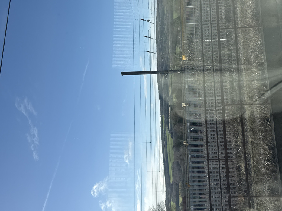 Captured through a window, a bright blue sky contrasts with the landscape beyond. A prominent contrail streaks across the sky. Parallel railway lines dominate the right side of the frame, running adjacent to a green, undulating terrain. Overhead power lines stretch across the scene, supported by dark poles. The image suggests a view from a moving train.