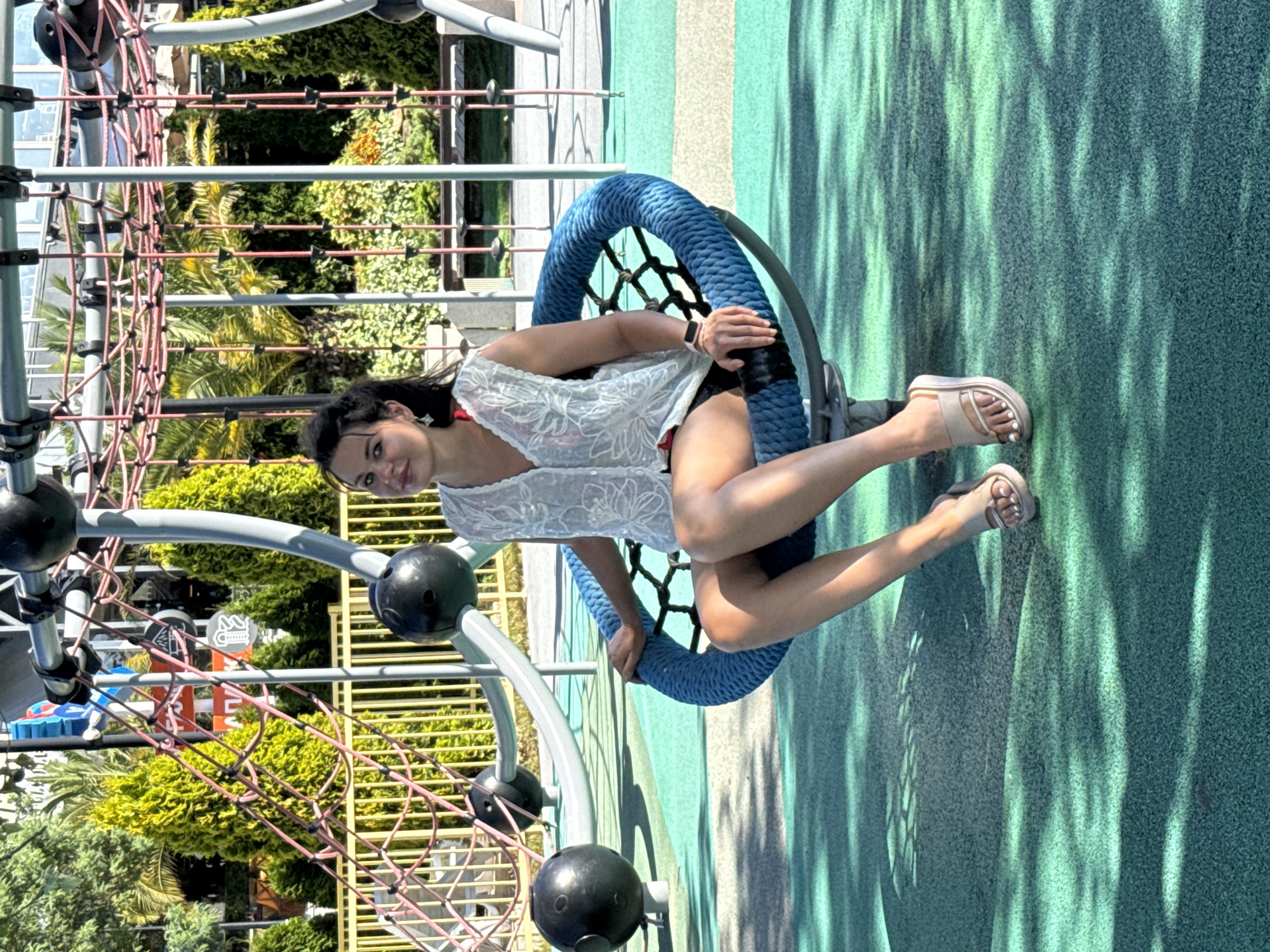 A young woman with dark hair pulled back in a ponytail sits in a blue rope swing chair on a green and turquoise playground surface. She is wearing a white sheer sleeveless top over a red garment and black shorts, and beige platform sandals. The playground equipment, including climbing nets and a slide, is visible in the background, along with lush green trees and palm trees. The scene appears to be outdoors on a sunny day. The environment suggests a public recreational area. The presence of palm trees and a vibrant, well-maintained playground points towards a location in a resort city. Given the context provided, this playground is situated in the city of Sochi, Russia, a well-known resort destination on the Black Sea coast.
