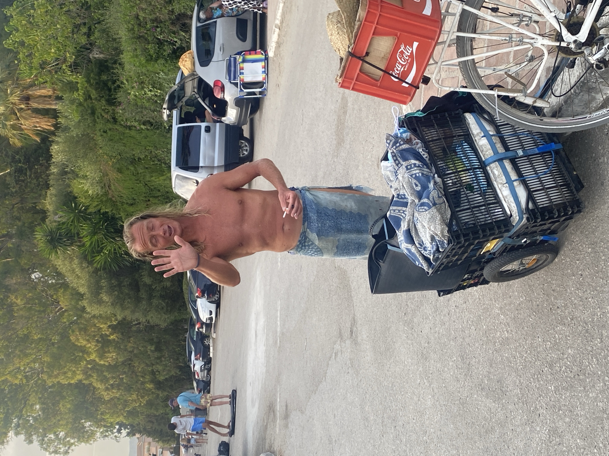 The image depicts an outdoor scene at Playa Del Sol-Villacana, Spain, likely a parking area near the beach. A shirtless man with long blonde hair, wearing a blue sarong and holding a cigarette, stands in the foreground facing the camera and waving with his left hand. He appears to be speaking. To his left, there is a black cart with two stacked crates, one filled with a blue and white patterned blanket and other items, and the other containing a mattress. To the right of the cart, a red Coca-Cola crate is visible, mounted on the back of a bicycle. In the background, several cars are parked, and lush green trees and foliage form a backdrop. Some people are seen in the distance near the cars. The ground is paved with asphalt.