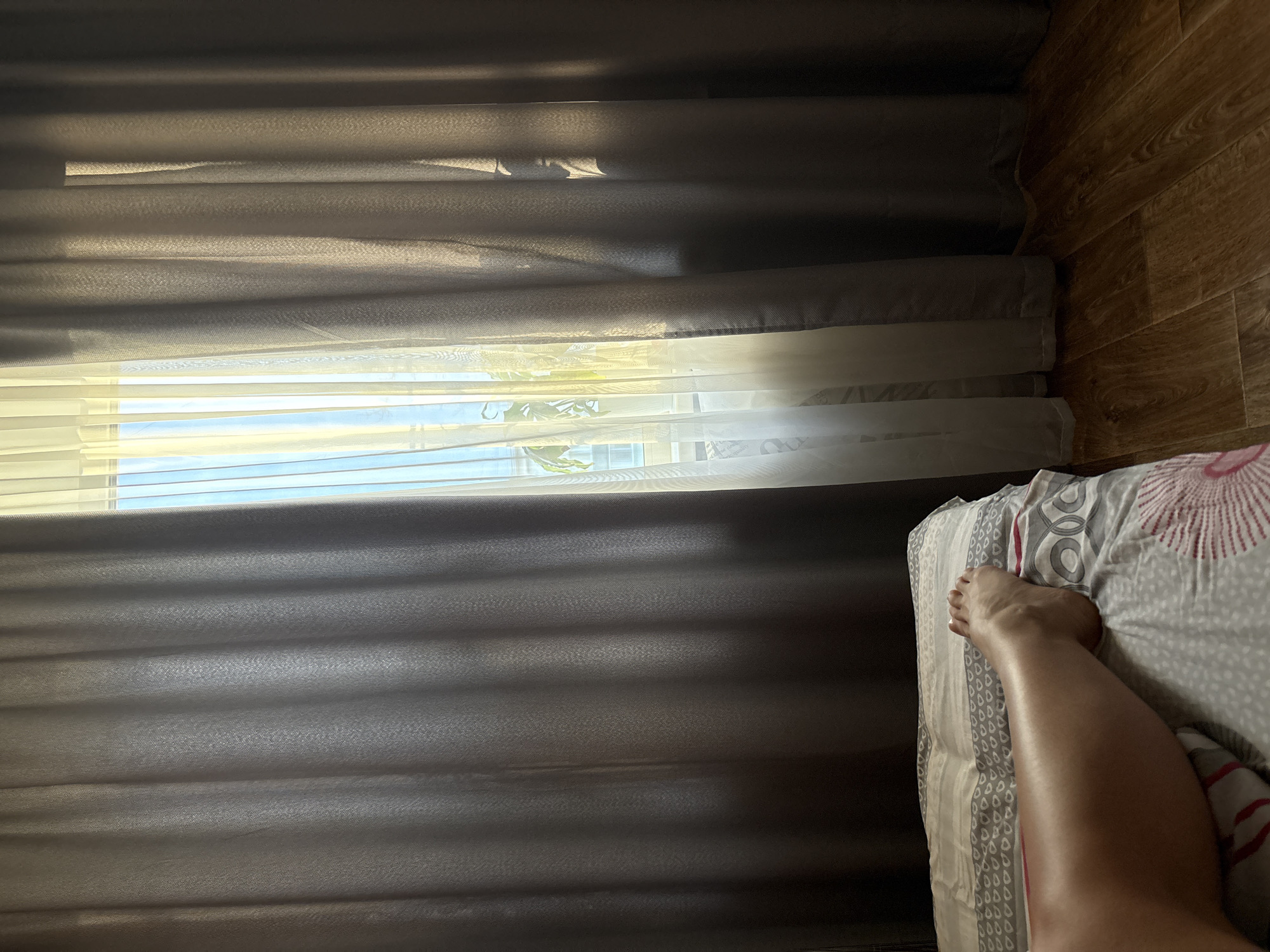 The photograph shows a bedroom with a window covered by gray curtains and blinds. Sunlight filters through the window. A portion of a bed is visible in the lower right corner, with a patterned bedsheet and a bare human leg resting on top. The floor is made of wood laminate. The location is unspecified and cannot be inferred from the image.