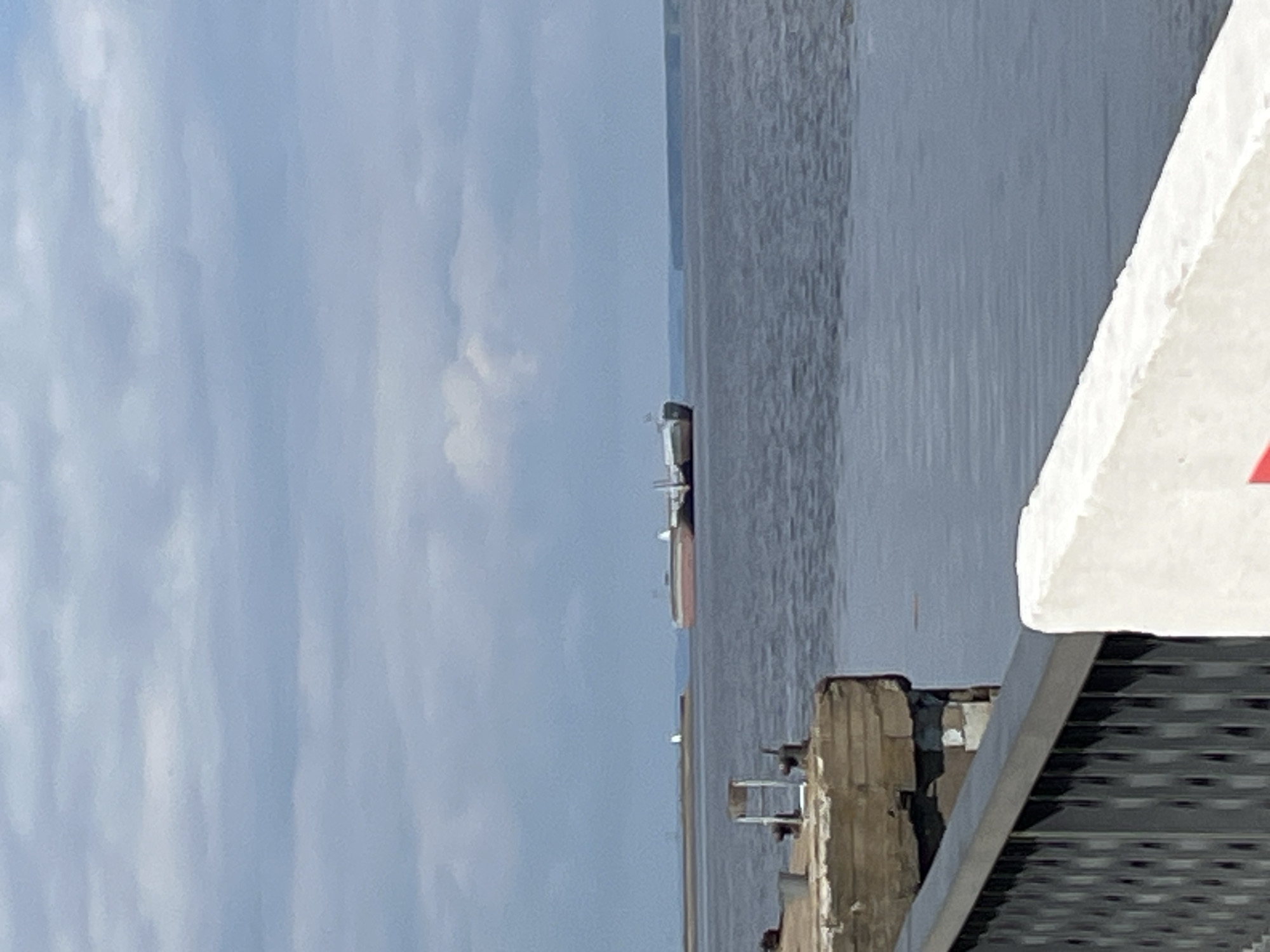 The image, taken in Chaikovsky, Russia, depicts a tranquil daytime scene along a wide body of water, likely a river or reservoir, under a partly cloudy sky. The perspective is from a raised position looking across the water.
In the immediate foreground on the right, a prominent white concrete structure with a diagonal edge is visible, possibly part of a pier, building, or large barrier. A small, indistinct red shape is faintly visible on its side. Beneath this concrete, a dark, textured surface, possibly a metal grate or patterned flooring, suggests part of a platform or walkway. To the left of these modern elements, an older, weathered wooden or concrete pier with visible railings or cleats extends into the water.
The expansive water surface, occupying the midground, appears calm with subtle reflections. In the distance on the water, several boats or barges are visible. One notable vessel has a distinct red-orange hull and a white superstructure, suggesting a cargo ship or a larger river transport vessel. Other, smaller, mostly white boats or barges are seen further along the horizon.
The background is dominated by a light blue sky filled with soft white and grey clouds, indicating an overcast or partly cloudy day. No people are visible, and there is no specific activity or event actively taking place, lending a quiet and serene quality to the scene. The overall impression is that of a functional riverfront or port area. No discernible text is visible.