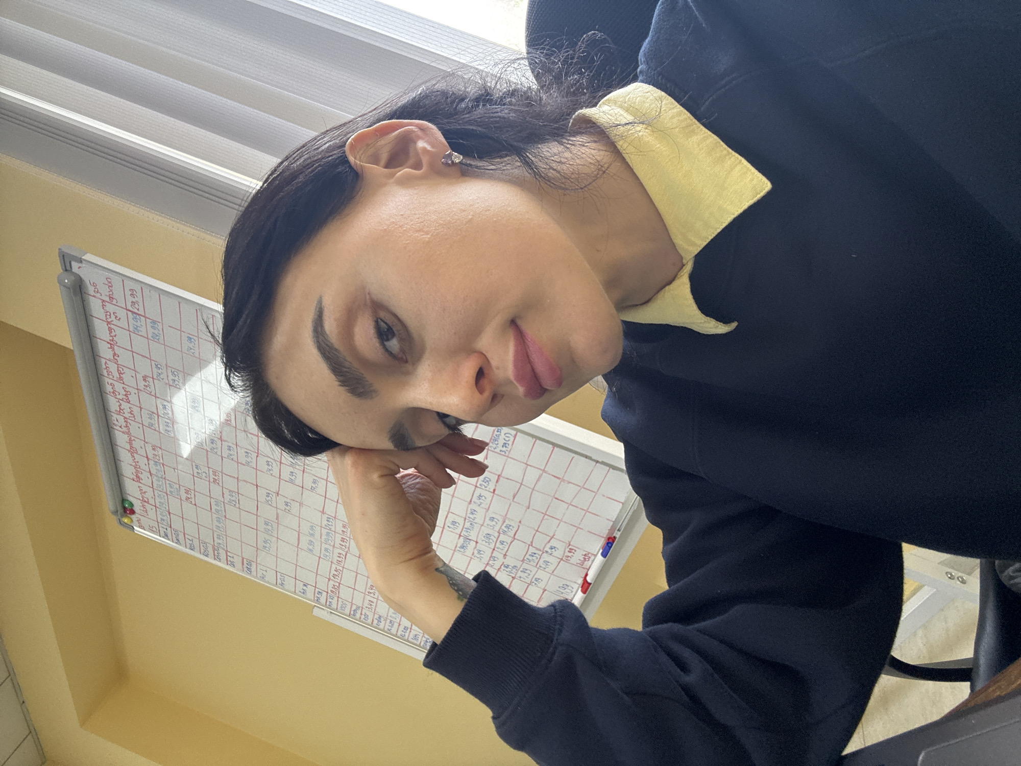 A person with dark hair, fair skin, and prominent eyebrows rests their cheek on their hand, with the arm in a dark blue sweater sleeve. A yellow collared shirt is visible beneath the sweater. The person is looking at the camera with a slightly playful expression, sticking out their tongue slightly. In the background, a white board with a grid of handwritten data is partially visible. Behind the whiteboard are partially visible white horizontal blinds. The wall appears to be painted light yellow. The OCR suggests the text on the board contains numeric data like "14,99 29,99 24,99." The OCR may contain the text "Tbilisi Georgia". The location is, therefore, possibly a business or school setting in Tbilisi, Georgia.