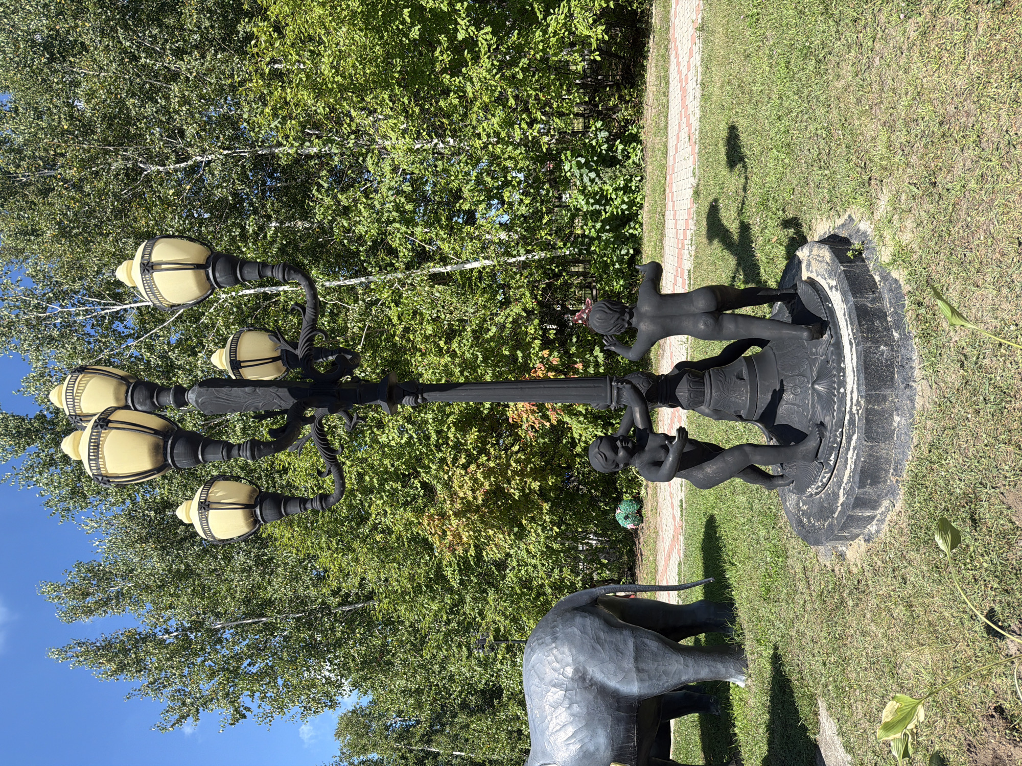 In Ivanovka, Russia, a black lamp post with five yellow lamps stands in a grassy area. Two dark-colored statues of nude children are positioned at the base of the lamp post, appearing to interact with it. To the left, a large, dark-colored statue of an elephant's hindquarters is visible. In the background, a dense line of green trees forms a backdrop, with a paved pathway made of red and white bricks running behind the statues. The sky is blue and clear.