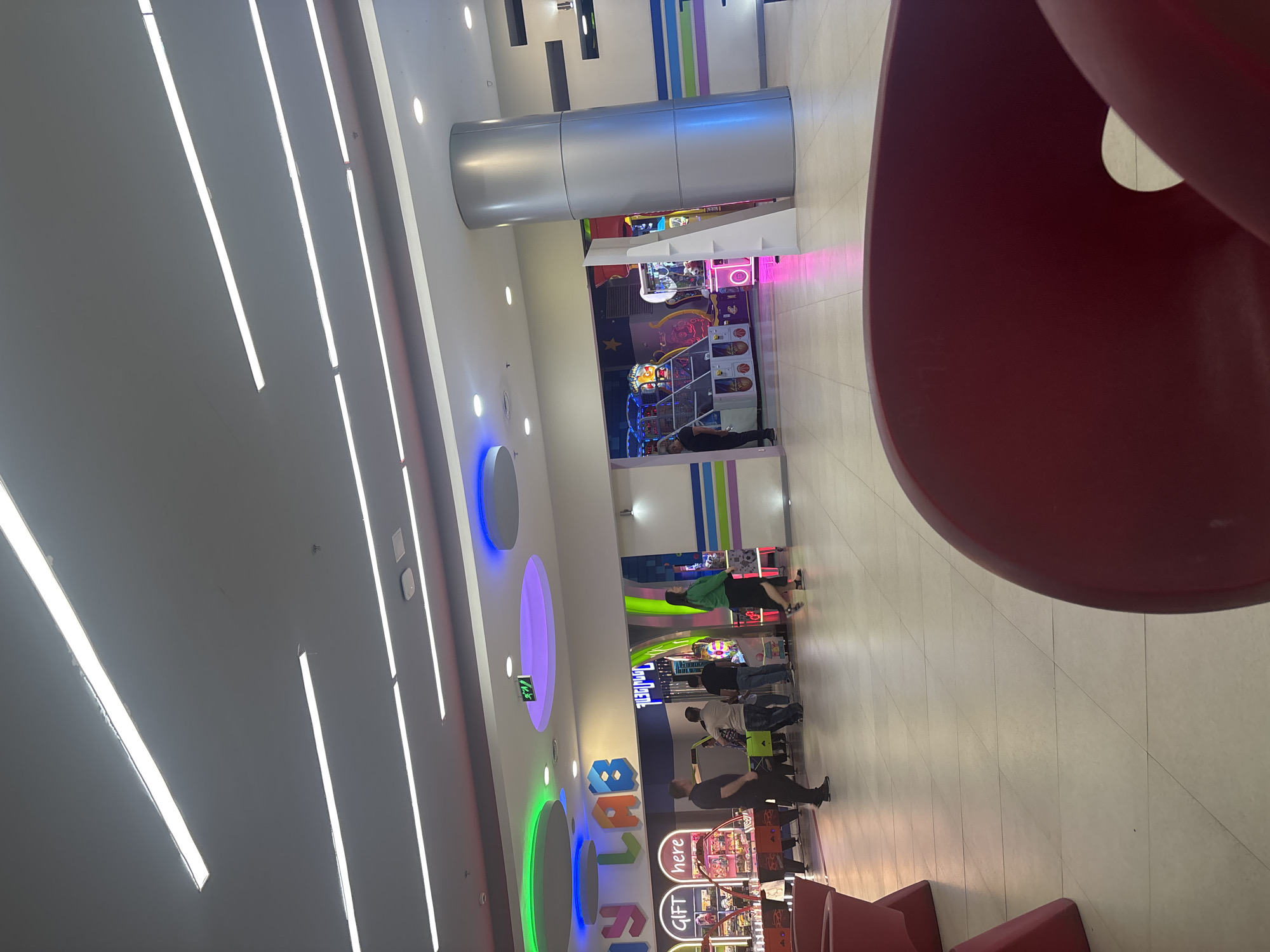 This image captures an indoor entertainment or gaming center, likely located in a mall or dedicated recreational facility. The space features a modern design with bright, linear white lights on the ceiling and colorful neon circular and linear lighting elements on the walls and structural pillars, in hues of blue, green, and purple.
Several individuals, predominantly adults, are visible throughout the area, engaged with various arcade machines. Activities include playing games, observing, and standing around the different stations. One person in a green shirt and black skirt is standing near an arcade machine in the background, while other groups are scattered across the floor interacting with the games.
Prominent features include a large, metallic silver cylindrical structure in the midground, possibly a decorative column or part of an architectural element. In the immediate foreground on the right, a large, curved, deep red structure, possibly part of a slide or an oversized seating arrangement, partially frames the scene. The floor is covered with light-colored square tiles.
Visible text includes "GIFT here" displayed on one of the arcade machines, and partially visible large, colorful letters that spell out "LABLAB" or "BABYLAB" on a wall to the left. The scene is well-lit, suggesting daytime or evening operating hours for the facility, portraying a lively and engaging atmosphere typical of an indoor entertainment venue in Yerevan, Armenia.