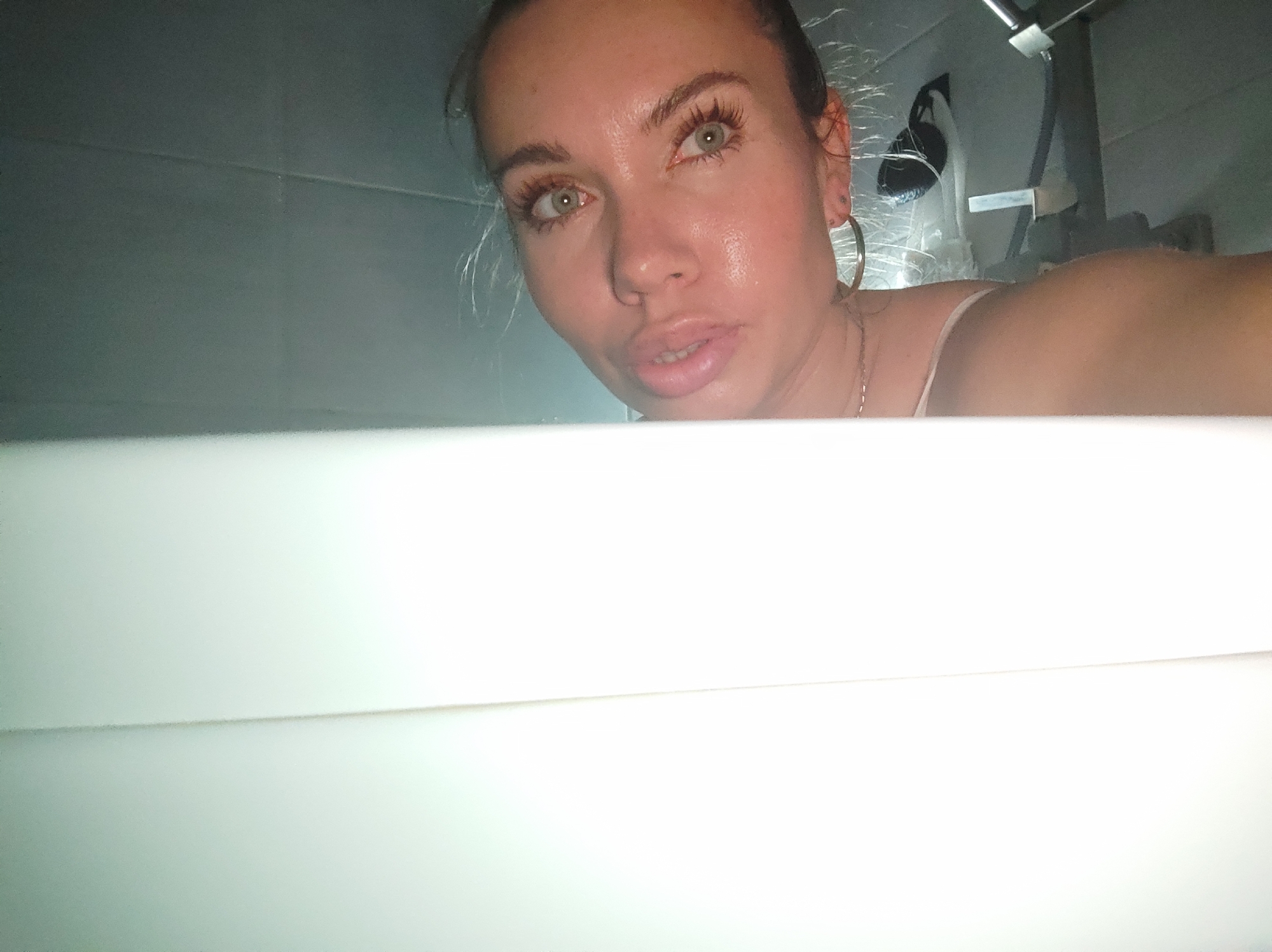 A woman with light skin, green eyes, and brown hair pulled back is looking directly at the camera. Her skin appears slightly wet or sweaty. She is wearing hoop earrings and a delicate necklace. The photo seems to be taken from a low angle, as a white, possibly rectangular object (likely a bathtub or a countertop) occupies the majority of the lower half of the frame.
In the background, there's a blurry view of what appears to be a bathroom setting. A chrome-colored fixture, possibly a showerhead or part of the plumbing, is visible, along with other indeterminate shapes suggesting standard bathroom equipment.
Based on the prompt, the location is presumed to be in Votkinsk, Russia.