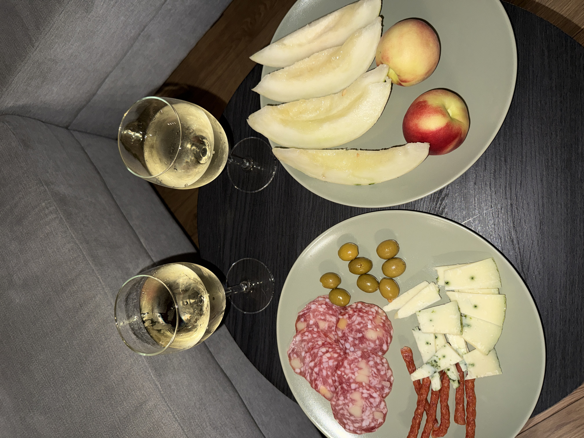 Two plates of charcuterie and fruit sit on a dark, textured table, next to two glasses of white wine. The plate on the left contains sliced salami, green olives, pieces of cheese with blue veins, and thin, reddish meat sticks. The plate on the right holds several slices of light-colored melon and two peaches. A grey sofa is visible in the background, partially obscuring the corner of a room. The lighting suggests an indoor setting, likely a home or restaurant. The image does not provide any specific information about the location in Szklarska Poręba, Poland.