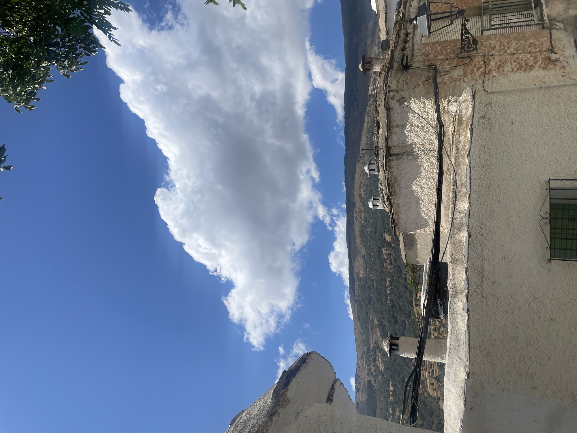 The image depicts a view of rooftops and a hillside in Capileira, Spain, under a bright blue sky with scattered white clouds. In the foreground, a white, textured wall dominates the left side, with another textured wall in the lower portion of the frame. Several chimneys with white caps are visible on the roofs of the buildings, which appear to be constructed with stone and plastered walls. Electrical wires are strung between the buildings. A black, ornate lamppost is mounted on the right side of a building, with a barred window below it. The background shows a densely vegetated hillside, with dry brush and scattered trees, leading up to a mountain range under the vast sky. A branch of a tree with green leaves is visible in the upper right corner of the image.