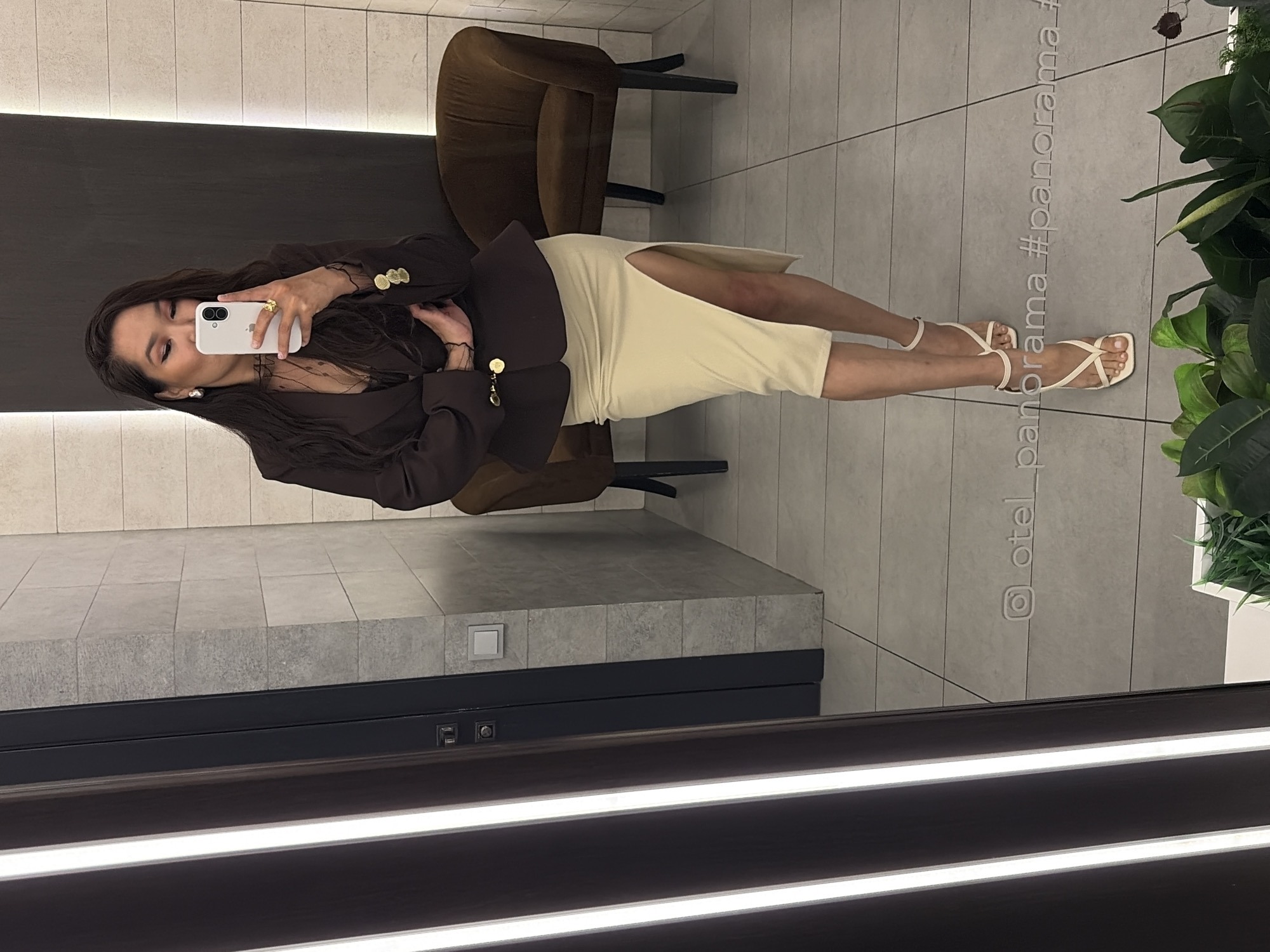 A full-length mirror selfie captures a woman posing in a modern indoor setting. The woman has long dark hair and is dressed in a dark brown blazer over a sheer top, paired with a light cream-colored midi-skirt featuring a high slit on her left leg. She is wearing small hoop earrings, a distinctive gold-toned ring, and light-colored strappy sandals. She holds a white smartphone (an iPhone) to take the photo, with her gaze directed towards the camera.
The background, visible as a reflection in the mirror, depicts a contemporary interior. A dark brown upholstered chair is positioned behind her. To the right, a white planter contains vibrant green leafy plants. The floor is covered with light grey square tiles. The bottom of the mirror frame incorporates two horizontal strips of bright white light, indicative of modern design.
Faintly visible text on the mirror surface reads "@otel_panorama #panorama_", strongly suggesting the location is a hotel named "Panorama." The overall atmosphere is sleek and modern, consistent with a hotel or upscale establishment. The image is presented with the subject oriented sideways, implying it was taken or uploaded in a rotated landscape format.