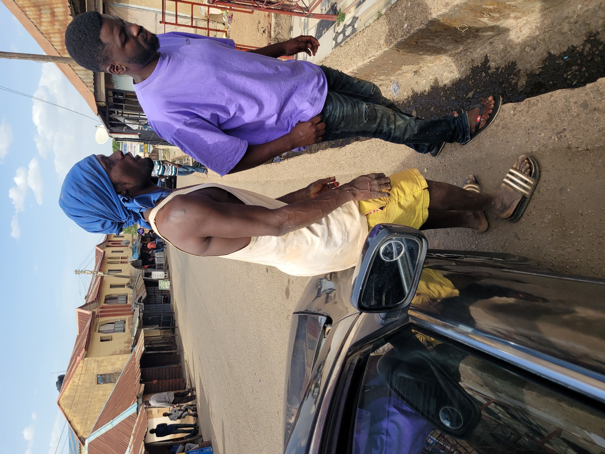 Two men are conversing beside a black car on what appears to be a street in Jos, Nigeria, under bright daylight with a clear blue sky.
The man closer to the camera, positioned on the left, has dark skin and a beard. He wears a light-colored sleeveless tank top, yellow shorts, and a blue headwrap. He has black and white striped slides on his feet. His hands are clasped at his waist.
Facing him, the second man, on the right, also dark-skinned with a beard, wears a purple short-sleeved t-shirt with faint white text "CALEM" visible across the chest. He has on dark, possibly ripped, jeans and black flip-flops. His hands are resting at his sides.
Between the men and the foreground is the side of a black car, possibly a sedan, with its side-view mirror prominent. Reflections on the car's surface show parts of the surroundings. The ground is a mix of concrete and dirt, with some uneven sections.
In the background, a street extends, flanked by multi-story buildings constructed with light-colored plaster and reddish-brown corrugated metal roofs. These structures appear to house shops or residences. One building displays a sign with "COMMUNITY DEVELOPMENT INITIATIVES" and "SALOON" visible. A small satellite dish is mounted on another building. The overall scene depicts a casual interaction in a lively urban or semi-urban setting.