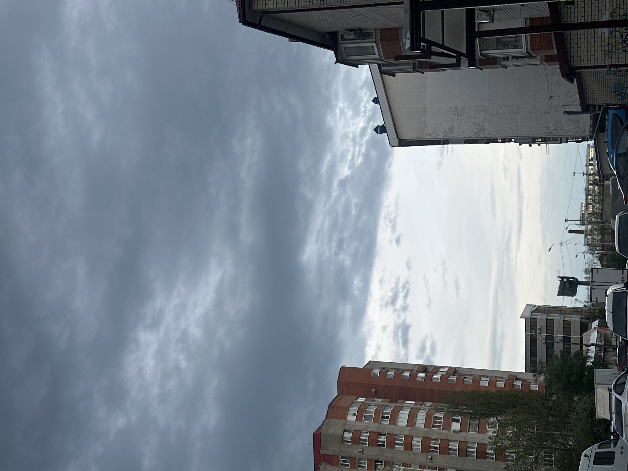 The image shows a street scene in an urban area, with buildings on either side and vehicles parked along the street. The sky is partially covered with dark, ominous clouds on the left, contrasting with a brighter, less cloudy area on the right. The building on the top of the image appears to be made of brick and white stone, while the one on the bottom is a brick building with multiple windows. There is a streetlight on the right and various objects and signs visible in the background.