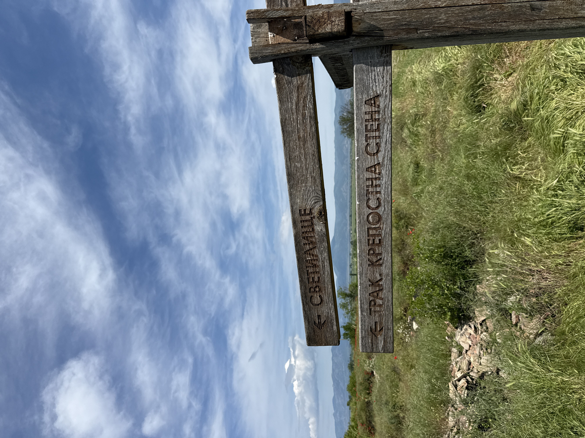 The image captures a wooden signpost located outdoors, with two hanging wooden signs indicating directions. On the left, the sign reads "СВЕТИЛИЩЕ" with an arrow pointing left, while the sign on the right reads "ТРАК. КРЕПОСТНА СТЕНА" with an arrow pointing left as well. The signposts are attached to a weathered wooden structure.
The background includes a cloudy blue sky in the top left, transitioning into a landscape with green grass, some scattered rocks, and possibly a view of a valley or lower terrain with vegetation and distant hills. There might be some red poppies visible in the grass near the right sign.
Based on the sign content and the query context, this image likely depicts a signpost directing towards archaeological or cultural sites near Kabile, Bulgaria, possibly indicating a sanctuary ("СВЕТИЛИЩЕ") and a Thracian fortress wall ("ТРАК. КРЕПОСТНА СТЕНА"). There are no people visible in the image.