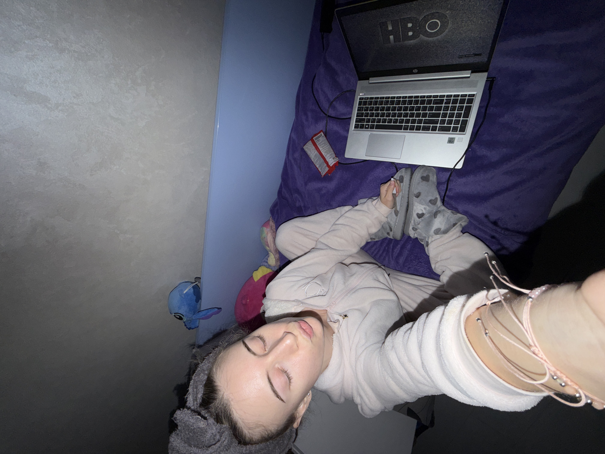 The image depicts a person lying on a bed in a room. The person is wearing a light pink pajama set and gray slippers with heart patterns. They have a gray towel wrapped around their head. A laptop displaying the HBO logo is positioned on the bed next to them, along with a red box of tissues. There are also several stuffed animals on the bed and a blue stuffed animal attached to the headboard. The location in Plovdiv, Bulgaria is not discernible from the provided image.