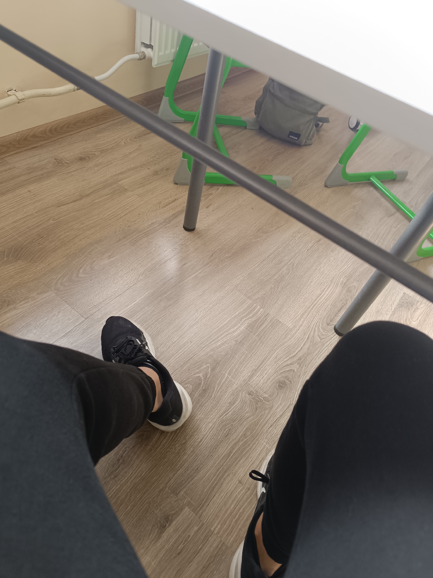 The image shows a first-person perspective of someone sitting at a desk. The person is wearing black leggings and black sneakers with white soles. The desk has a white top and a green and gray metal frame. The floor is made of light brown wood-look laminate. In the background, a gray Adidas backpack is visible under the desk, and a white radiator and pipes are visible against a beige wall.
It is not possible to verify this photo's exact location in Pleven, Bulgaria.