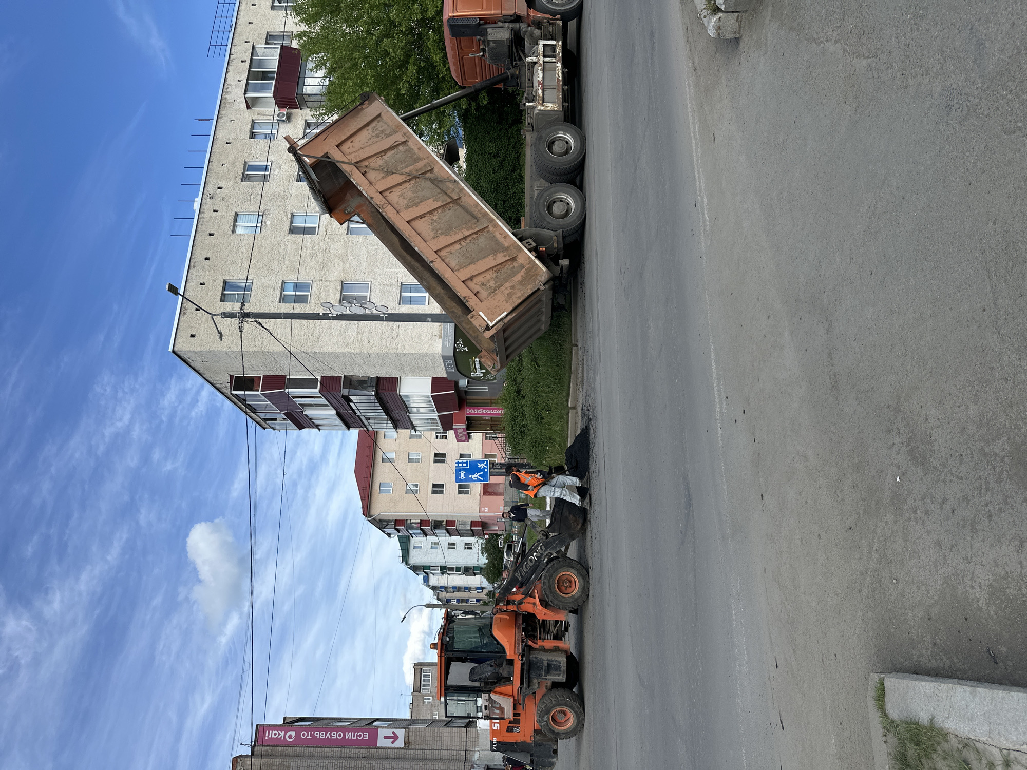 The image captures a street-level view of road construction in an urban environment. A large orange dump truck with its bed raised is in the process of unloading asphalt onto the road. Next to it, an orange Yigong 7118 loader is positioned to move the asphalt. Two workers wearing light-colored clothing and one worker in an orange vest can be seen near the machinery.
In the background, a multi-story building with gray and white facade and red accents is visible, along with green trees and a partially visible blue sky with scattered clouds. Street signs are affixed to a pole near the workers. Another sign is seen on the lower left, with Russian text. The road is asphalt, and the weather appears to be clear.