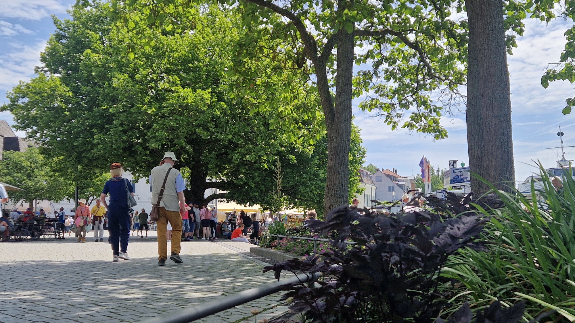 The image captures a sunny day in the city of Überlingen, Germany. In the foreground, a paved walkway leads towards a park-like area with large, mature trees casting shadows. Two individuals, a man and a woman, are walking away from the camera on the walkway. The woman is wearing a blue jumpsuit and a brown hat, and the man is wearing a light-colored vest over a blue shirt and khaki pants, also with a light-colored hat. Further into the scene, a variety of people are visible, some sitting at outdoor tables, while others are standing and conversing. In the background, buildings typical of a German town can be seen, along with some flags and a boat, suggesting a proximity to a harbor or waterfront. The overall atmosphere is relaxed and suggests a public gathering or leisure activity.