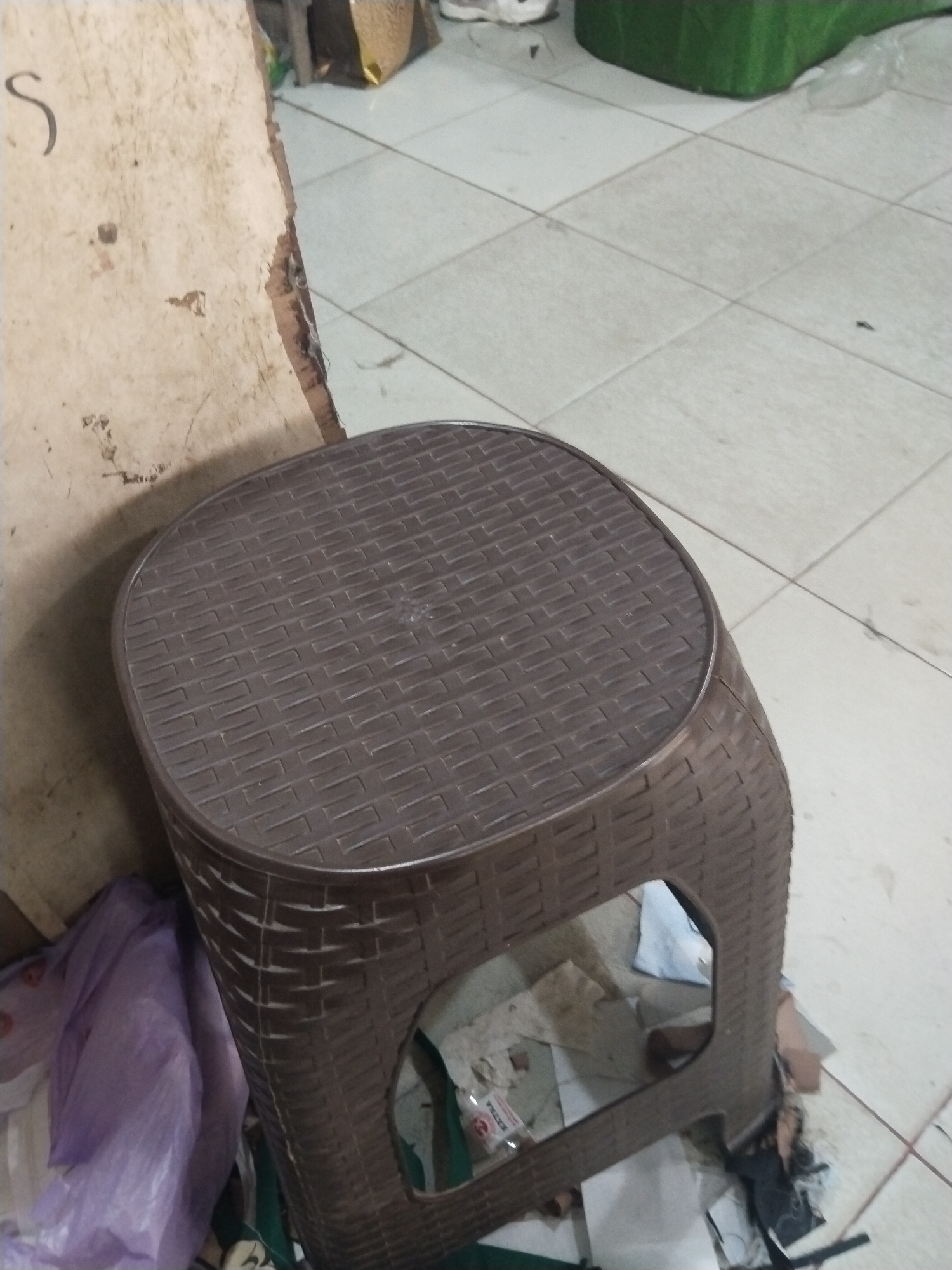 The image shows a brown plastic stool with a woven pattern on its surface. The stool has a square top and cylindrical base with an opening on one side. It is placed on a tiled floor. Next to the stool, there is a piece of a light-colored wall with a dark stain and a curved dark line, possibly a letter or a scratch. To the left of the stool, a crumpled purple plastic bag is visible, along with other debris on the floor. In the background, a portion of a green object, possibly a piece of furniture or a container, can be seen. The overall setting appears to be indoors, possibly a storage area or a less-maintained part of a building, given the presence of scattered items and the condition of the wall. The floor is made of light-colored square tiles.