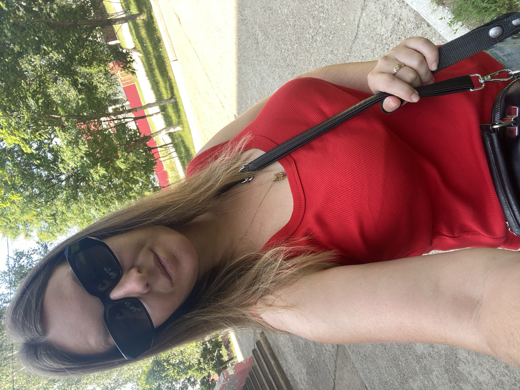 A woman wearing black sunglasses and a red sleeveless top poses for a selfie outdoors. She has blonde hair and is wearing a gold necklace and a ring on her right hand. A black purse with silver hardware is slung across her body. The background shows trees, grass, a paved area, and a red building with a yellow fence. The image appears to have been taken on a sunny day in Noviy, Russia.