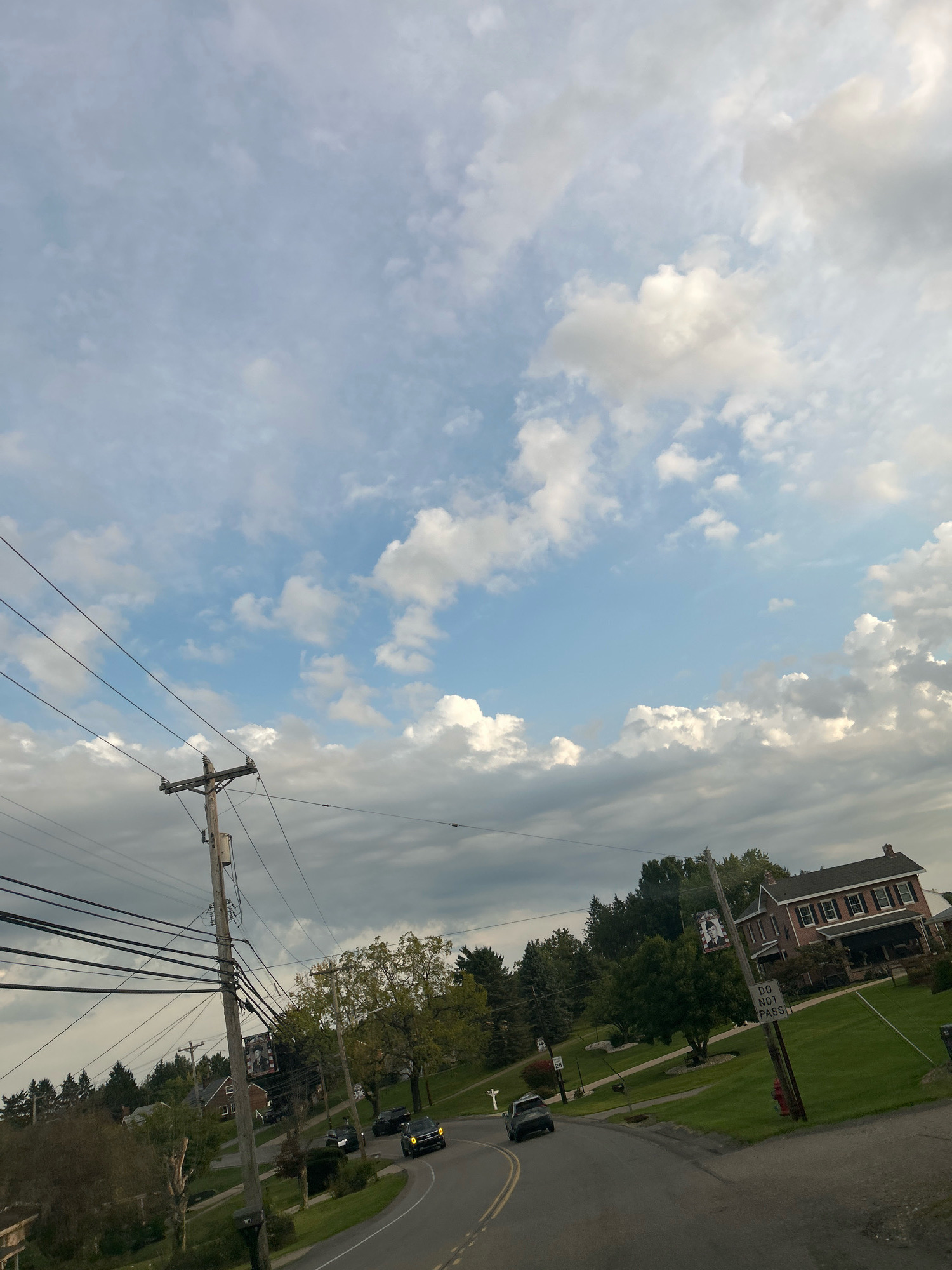 The image depicts a street scene in Robinson Township, United States of America. The perspective is from a vehicle moving along a paved road that curves to the right. On the left side of the road, a utility pole with numerous wires is prominent. Across the street, a large brick house with a dark roof, multiple windows, and a covered porch stands on a grassy incline. Adjacent to the house, a smaller wooden sign reading "DO NOT PASS" is mounted on a pole. Several trees are visible, some with green foliage and others appearing bare. Three cars are present on the road; one is a black SUV, another is a dark sedan, and a third appears to be a police car. The sky is partially cloudy with patches of blue visible. The overall environment suggests a rural or suburban setting.
