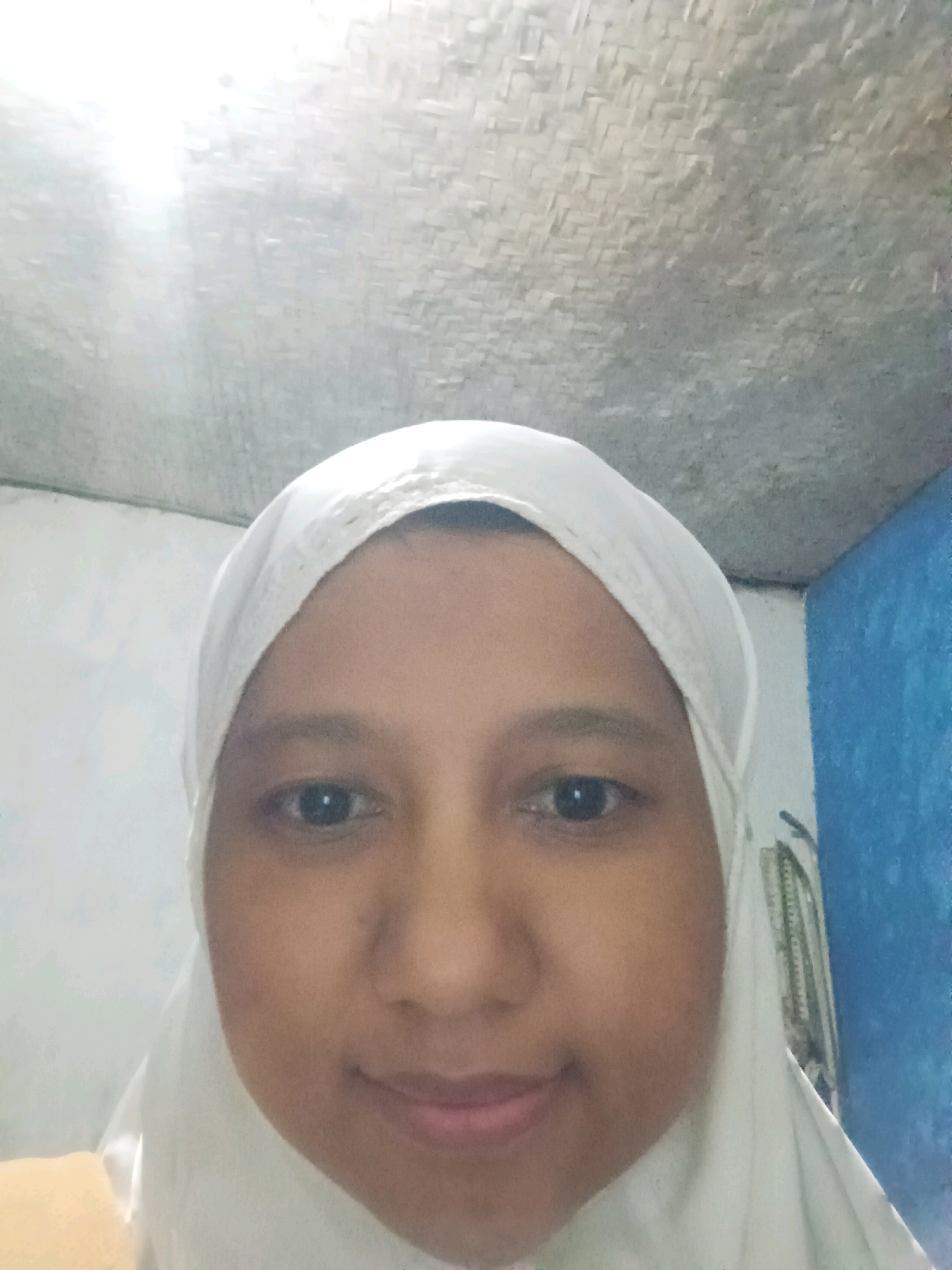 A close-up selfie of a young person wearing a white hijab is captured indoors. The subject is looking directly at the camera, with their face occupying most of the frame. The background is dimly lit, showing a textured ceiling and a blue wall on the right side. The overall lighting is bright, with a strong light source coming from the upper left, creating a slight glare in the image.