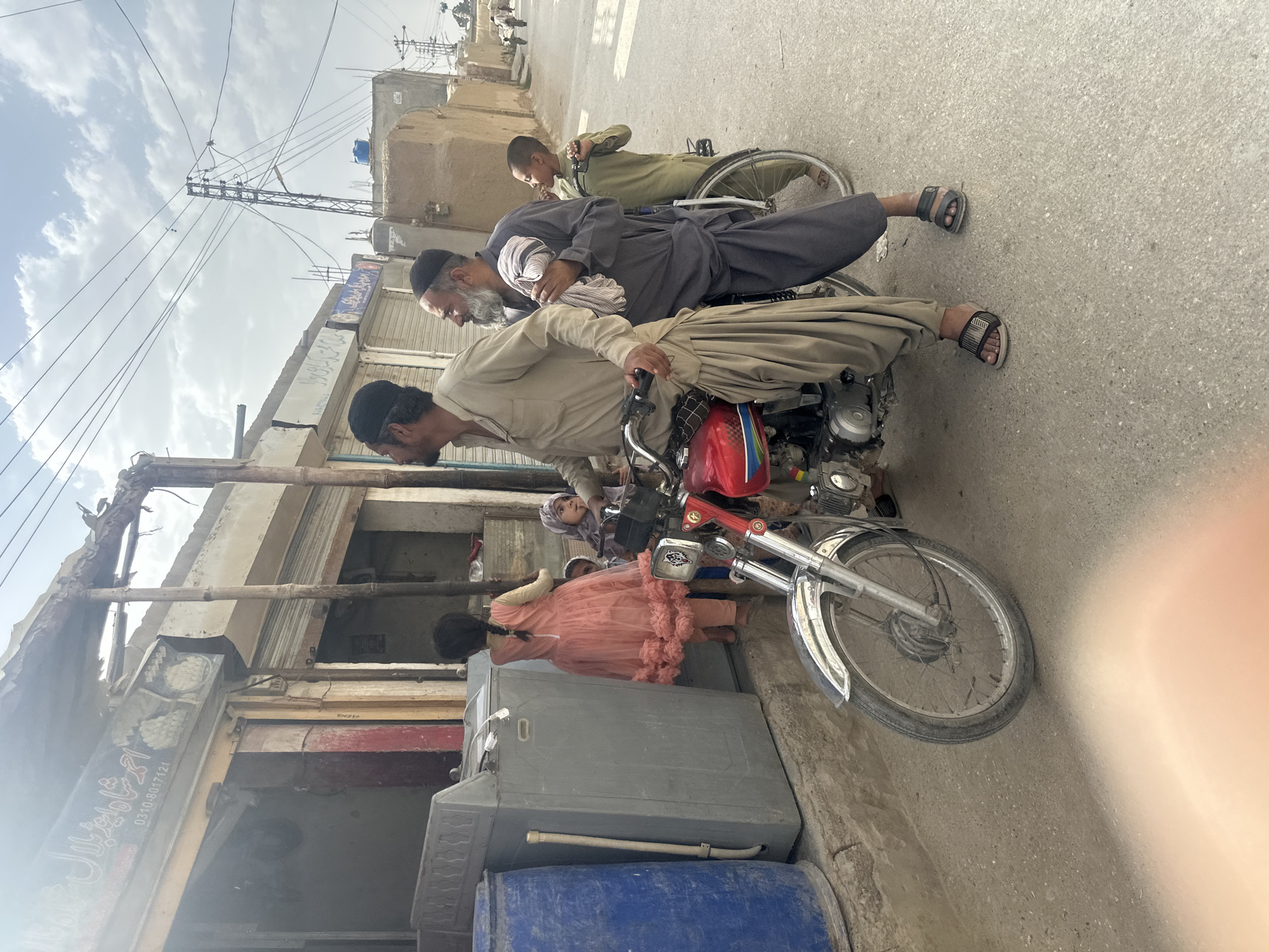 A street scene in Quetta City Tehsil, Pakistan shows three men and two girls on a street. One man is sitting on a motorcycle. Another man is riding in a wheelchair. A boy stands near the man in the wheelchair. Two girls are near the motorcycle. The street scene includes buildings with damaged exteriors. A washing machine and a blue barrel stand on the street near the motorcycle.