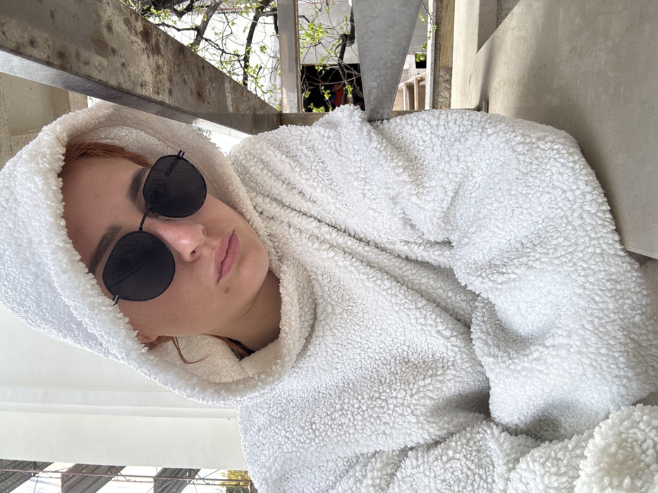 A female individual is depicted in a close-up, chest-up shot. She wears a cream-colored, textured "teddy fleece" hooded sweatshirt, with the hood pulled over her reddish-brown hair. Dark, circular-frame sunglasses obscure her eyes. Her expression is neutral, with closed lips, and a noticeable mole is present on her upper left cheek. She is positioned outdoors, leaning on a light-colored concrete surface. To her left, a white or light-colored building element is visible, with construction scaffolding and wooden planks in the background. To her right, a gray metal railing or structural beam runs diagonally, with concrete steps and sparse tree branches displaying light green leaves in the background. The scene is illuminated by natural daylight. The precise location within Budva, Montenegro, is not discernible from the visual information.