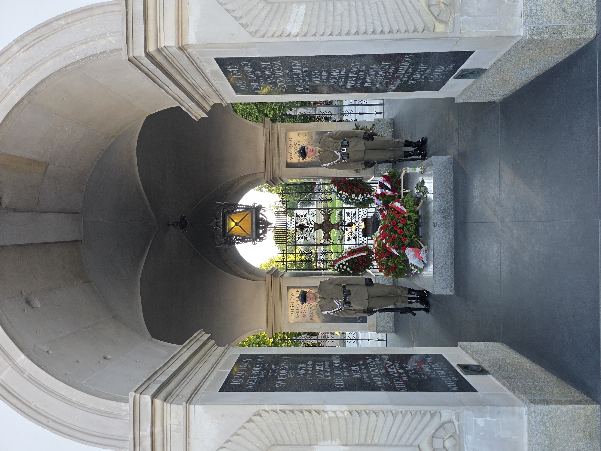 The image shows the Tomb of the Unknown Soldier in Warsaw, Poland. Two uniformed soldiers stand guard on either side of a tomb, which is adorned with floral wreaths. The Tomb of the Unknown Soldier is an iconic memorial located in Piłsudski Square, Warsaw, Poland, commemorating the soldiers who have died in defense of their country. The architectural style of the monument, with its grand arches and classical columns, is evident in the background. The soldiers are dressed in military uniforms, standing at attention with rifles. The environment appears to be outdoors, with natural light suggesting daytime. The overall atmosphere is solemn and respectful, as expected at a war memorial.