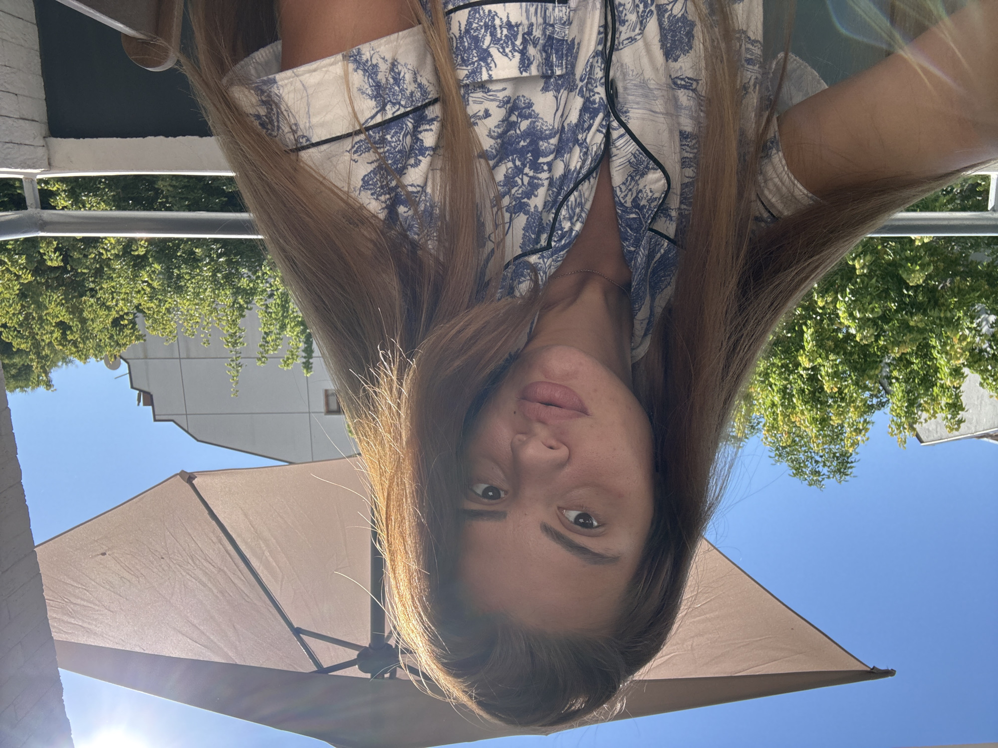 A young Caucasian woman with long, brown hair is captured in a selfie, taken outdoors on a balcony. She is wearing a white short-sleeved pajama top with a blue floral pattern. The background reveals a clear blue sky, a large light brown umbrella providing shade, lush green trees, and parts of buildings. The sun is shining brightly, casting a lens flare into the image. The scene is likely a residential balcony in Plovdiv, Bulgaria, given the architectural style and vegetation typical of the region.