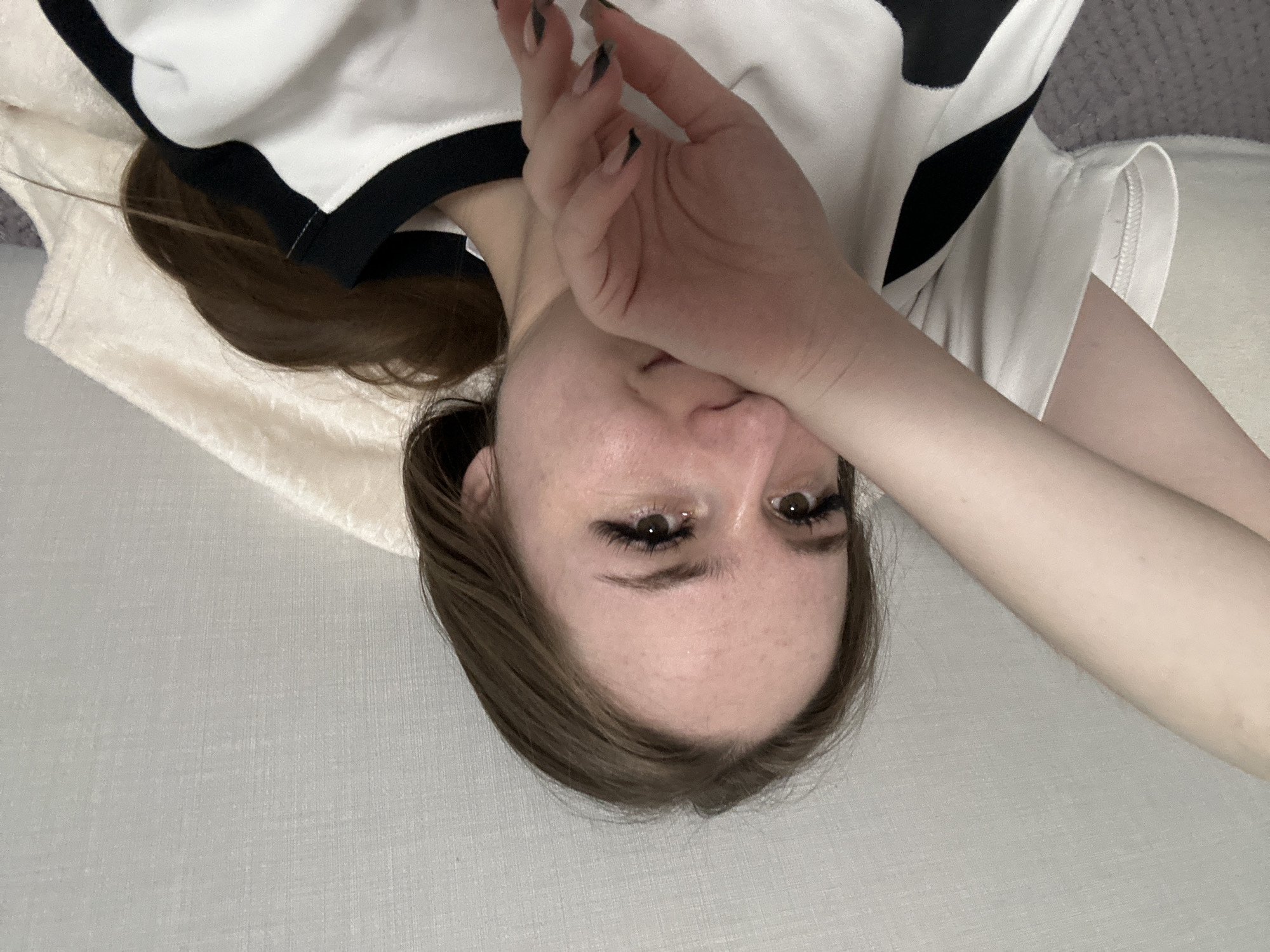 A young woman is depicted in a close-up, rotated portrait, appearing to be lying down. Her head is positioned towards the bottom of the frame, with her body extending upwards. She has medium brown hair, which is casually spread around her head and shoulders. Her eyes, which are open and brown, look directly at the viewer, featuring prominent, dark eyelashes, possibly enhanced with mascara or extensions. Light freckles are visible across her forehead.
Her right hand covers the lower part of her face, from her nose down to her chin, with her fingers resting gently across her mouth. The nails on her visible fingers are long and shaped, featuring dark tips, possibly a black or dark grey French tip style. She is wearing a white top with distinct black trim or a black pattern around the neckline and shoulder area.
The setting appears to be indoors, likely a bedroom or a comfortable lounge area. She is resting on a light grey, subtly textured fabric, which covers the majority of the lower and right portions of the image. Another softer, light beige or off-white textured fabric, resembling a blanket or pillow, is visible underneath her head and upper body. The overall impression is a relaxed, personal moment captured in a direct gaze despite the partially obscured face. No text is visible in the image.