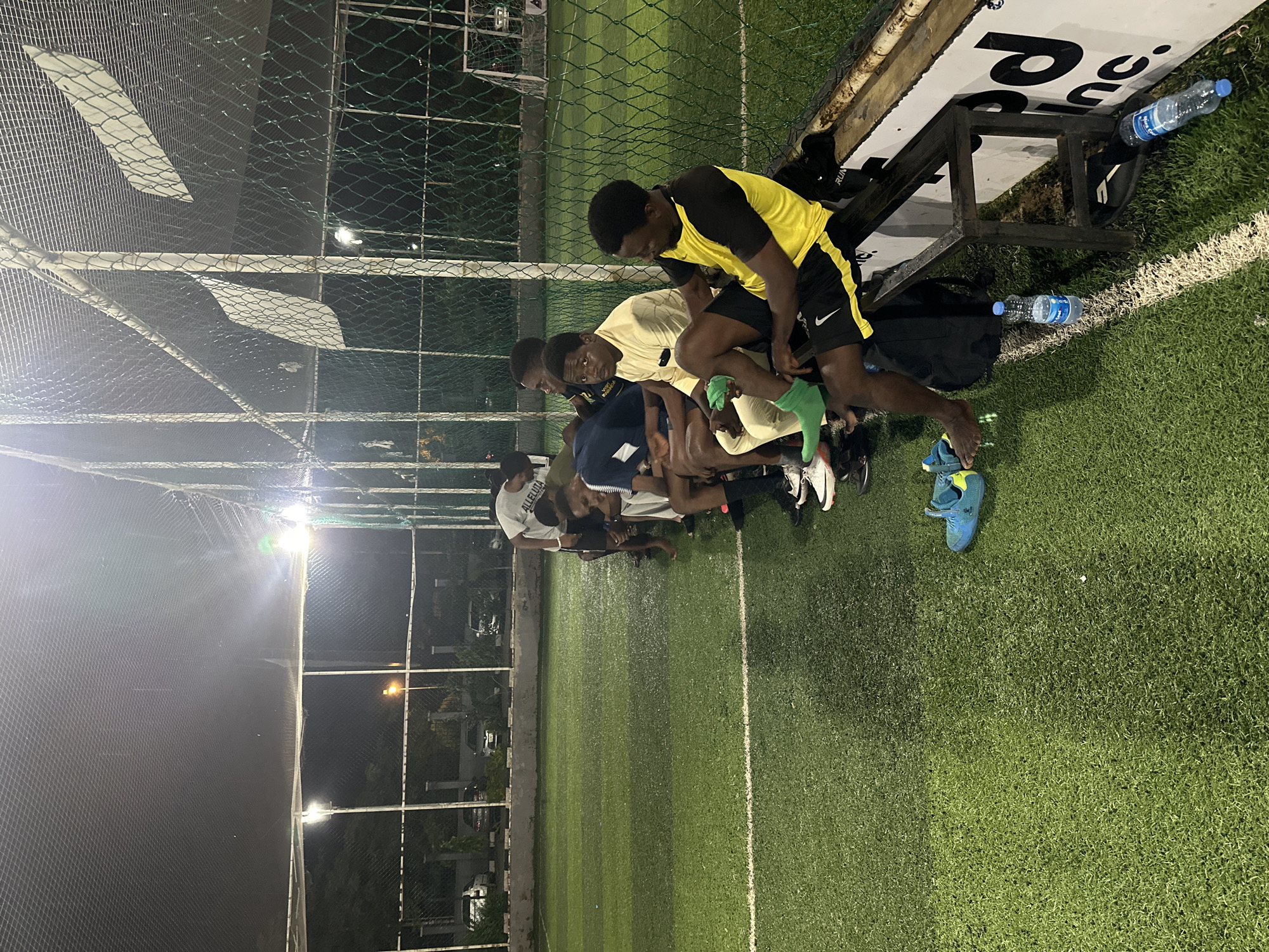 A group of young men, likely soccer players, are sitting on a metal bench along the side of a lit artificial turf soccer field at night. The field is green with white boundary lines. The players are casually dressed, some in athletic wear, and appear to be resting or waiting. One individual in the foreground is wearing a yellow and black soccer jersey and black shorts, and is seen adjusting green socks. Another person to his left is wearing a light-colored long-sleeved shirt and white pants, and is looking forward. Behind them, other individuals are also seated or standing. Water bottles are visible on the ground near the bench. In the background, a chain-link fence encloses the field, and beyond that, there are some trees, parked cars, and buildings, suggesting an urban setting within Abuja, Nigeria. The overall lighting is artificial, from overhead floodlights.