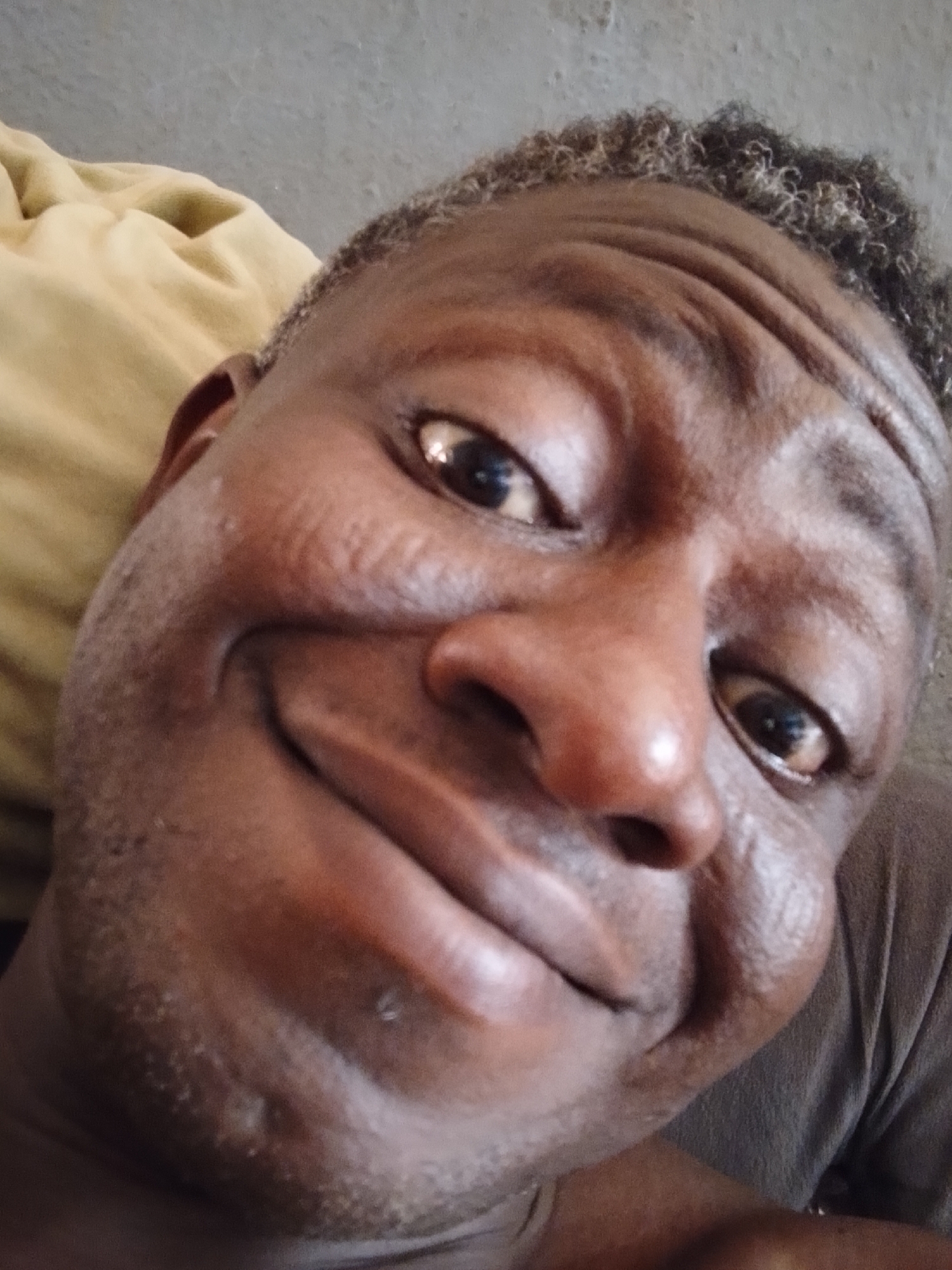 A close-up, selfie-style photograph depicts a middle-aged Black man. He is smiling broadly, with his head tilted slightly. His eyes are dark brown, and his hair is short, graying, and curly. Wrinkles are visible on his forehead and around his eyes. The background appears to be an indoor setting, with a beige pillow or cushion visible on the left side. The man is wearing a dark brown shirt or t-shirt. The location and the city are unknown.