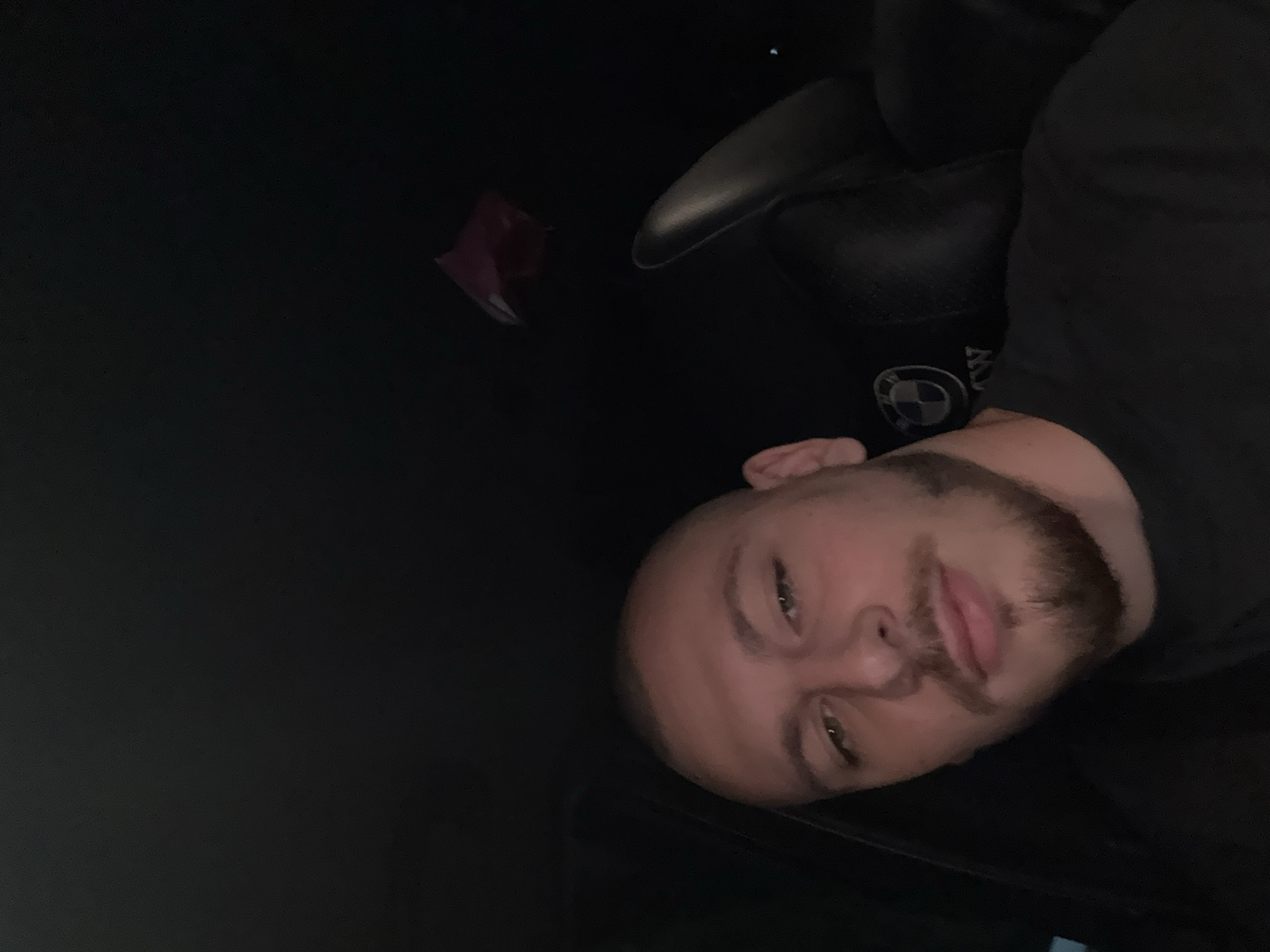 In a dimly lit car, a man with a shaved head and a beard is seen from the chest up, looking directly at the camera. He is wearing a black t-shirt, and the headrest of the car seat behind him features a BMW logo. The interior of the car is dark, with a small, reddish object visible in the upper right portion of the frame, possibly a piece of clothing or accessory. The overall impression is a candid, possibly self-taken, photograph within a vehicle at night. There is no indication of the location being Stara Zagora, Bulgaria.