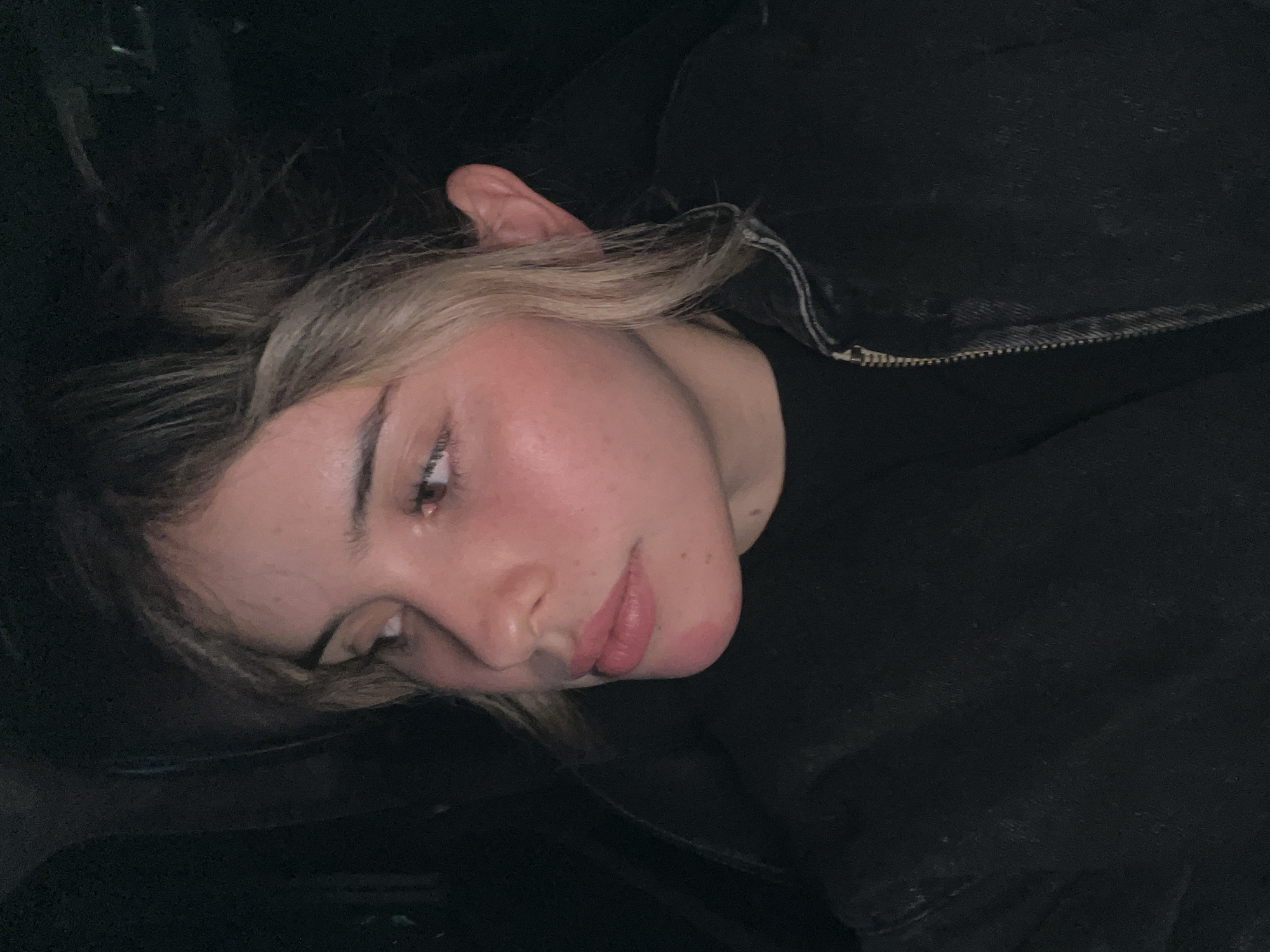 A close-up, head-and-shoulders shot captures a young woman's profile as she looks to her left. Her hair, a blend of dark brown and blonde, is partially styled with a braid. Her skin has a flushed appearance, and she is wearing a dark jacket with a zipper. The background is dark, suggesting an indoor or nighttime setting. The image was taken in San Fernando del Valle de Catamarca, Argentina.